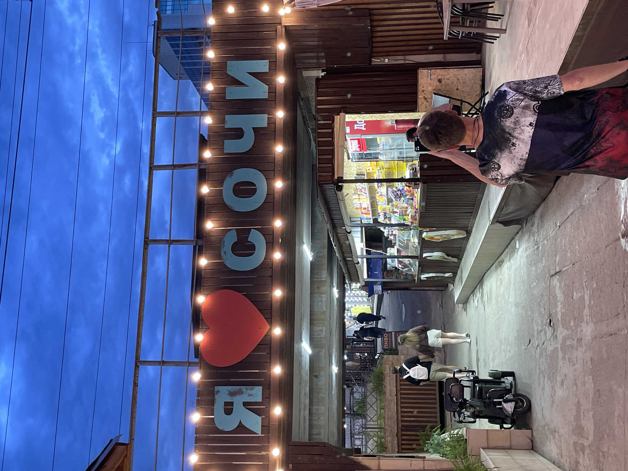 The image depicts an outdoor scene in Sochi, Russia, at dusk. A wooden structure with the word "ИЗООB" in blue Cyrillic letters and a red heart symbol is illuminated by strings of lights. A man wearing a patterned shirt and red shorts stands to the side, seemingly looking at or interacting with a storefront within the structure. Several people are walking in the alleyway, and a mobility scooter is parked nearby. Small shops line the alleyway. The sky above is a deep blue, indicating twilight.
