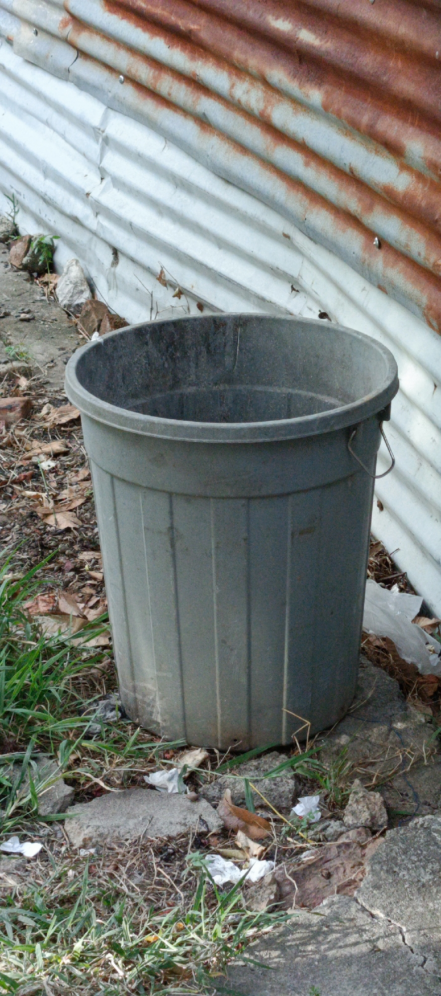 A weathered, dark gray plastic trash bin sits on uneven, rocky ground covered with dry leaves and sparse green grass. The bin is cylindrical with vertical ribbed detailing and a reinforced rim. A metal wire handle is attached to one side. In the background, a corrugated metal wall, partially rusted and weathered, stands behind the bin. Small pieces of white plastic and other debris are scattered around the base of the bin and on the ground. The overall scene suggests an outdoor, possibly neglected, area. This location is identified as Pashiebeutong, Indonesia.