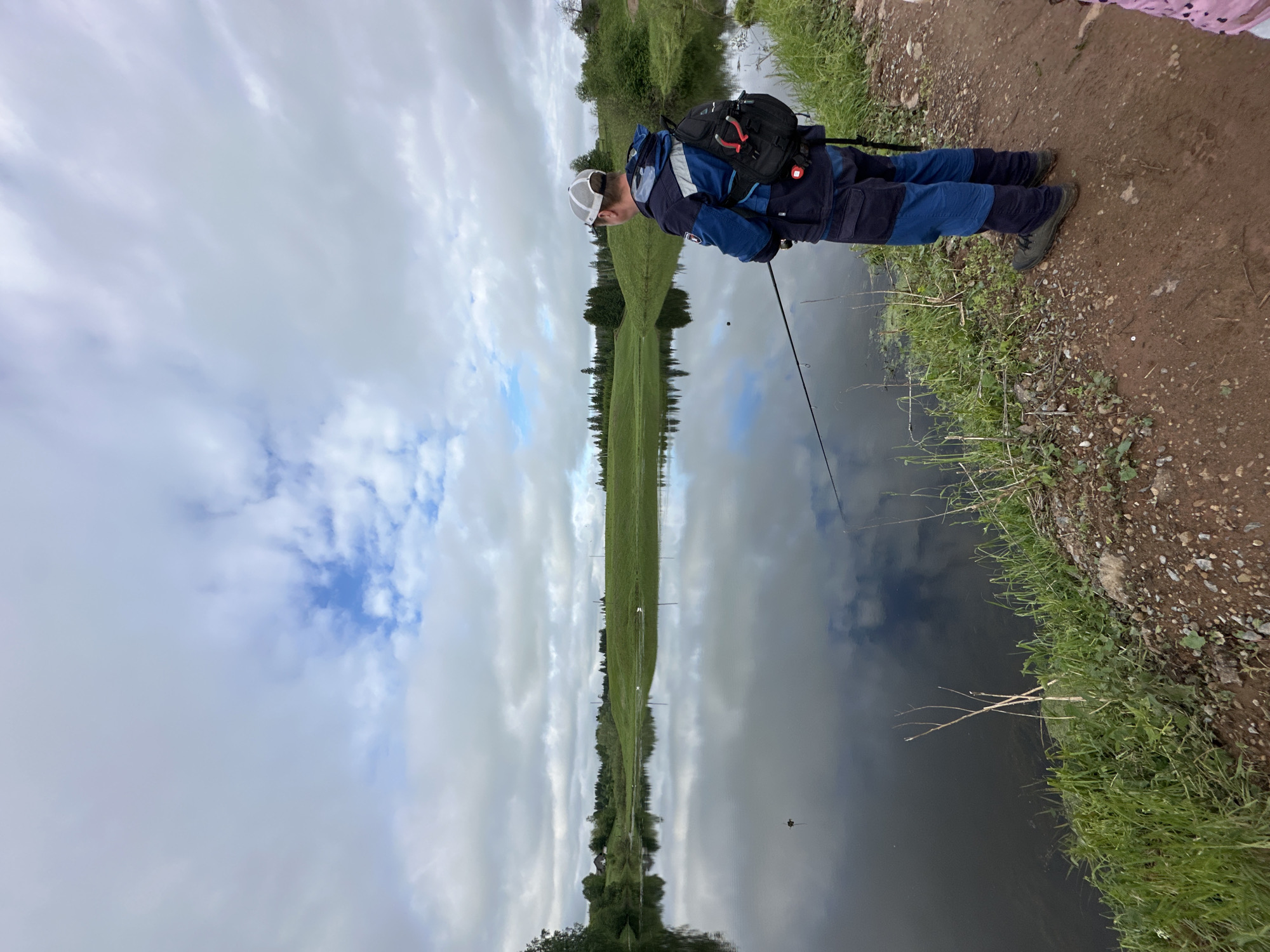 A person is fishing by a body of water. The scene depicts a person wearing a dark blue jacket, trousers and a cap, along with a black backpack, standing near the edge of what appears to be a lake or wide river. They are holding a fishing rod, with the line extended into the water. The water reflects the sky, which is filled with clouds. The opposite bank of the river is visible, showing a grassy field. The edge where the person is standing is covered with green grass and a dirt path. While I have visual information, I do not have information on geographic locations.
