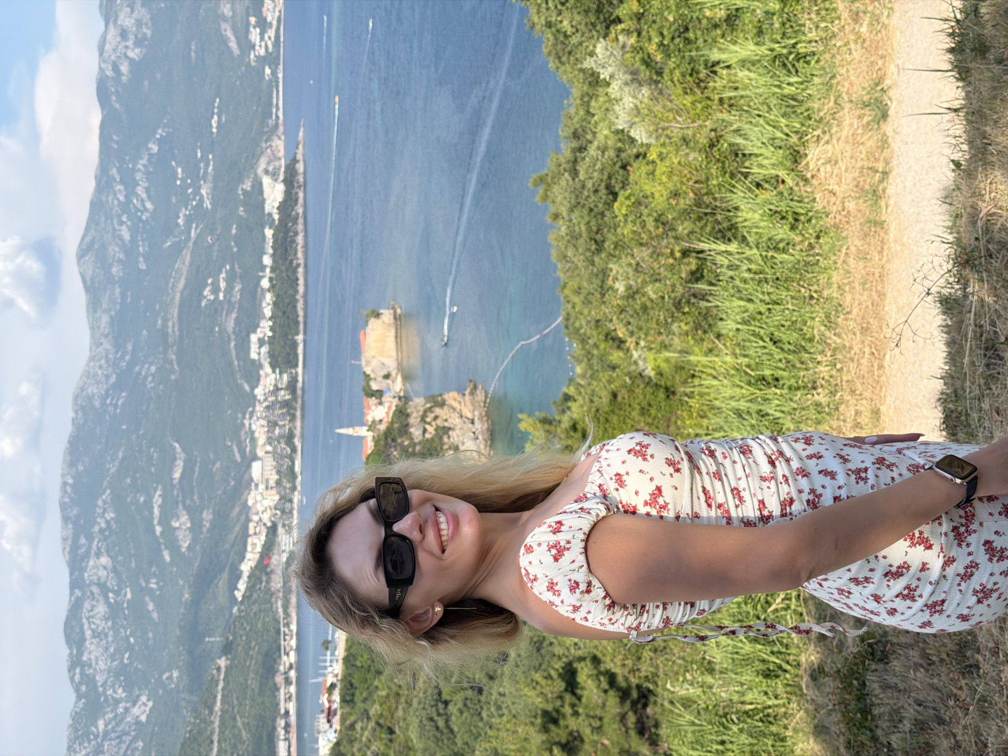 A woman is standing in front of a scenic view of the Adriatic Sea and the city of Budva, Montenegro. The woman is smiling and looking at the camera. She is wearing a floral print dress and sunglasses. In the background, there are mountains, the sea, and the city with its historic Old Town and a church spire. Several boats are visible on the water. The foreground consists of green vegetation and a dirt path.