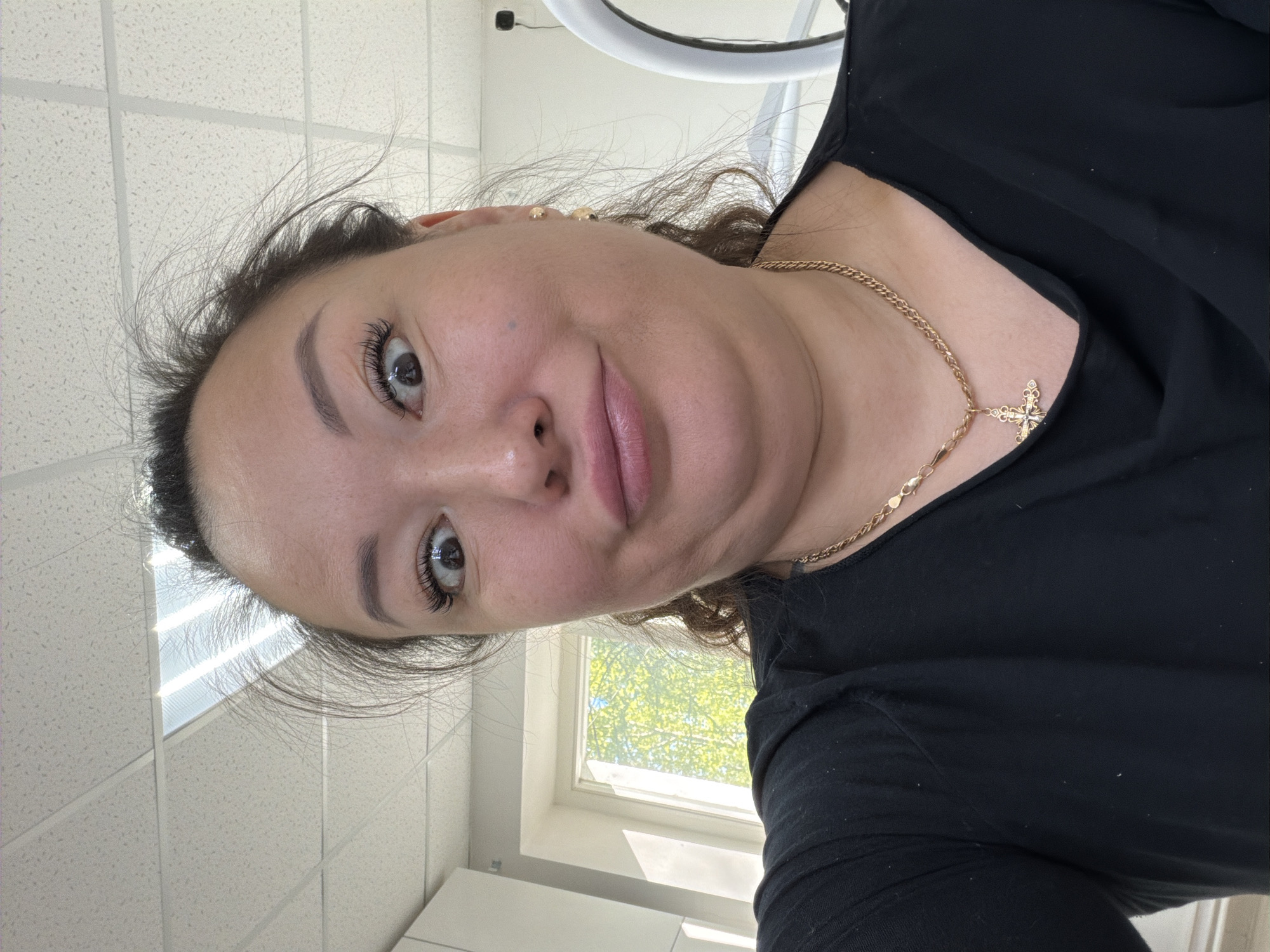 A woman is the primary subject of this vertically oriented close-up photograph. She has brown eyes and dark hair pulled back from her face, and is wearing eyelash extensions and a gold necklace with a cross pendant. The woman is wearing a black top. The photograph appears to have been taken indoors, given the visible ceiling tiles, window, and other fixtures. The window reveals the presence of green trees outside. According to metadata, the image was taken in the city of Chaikovsky, Russia.