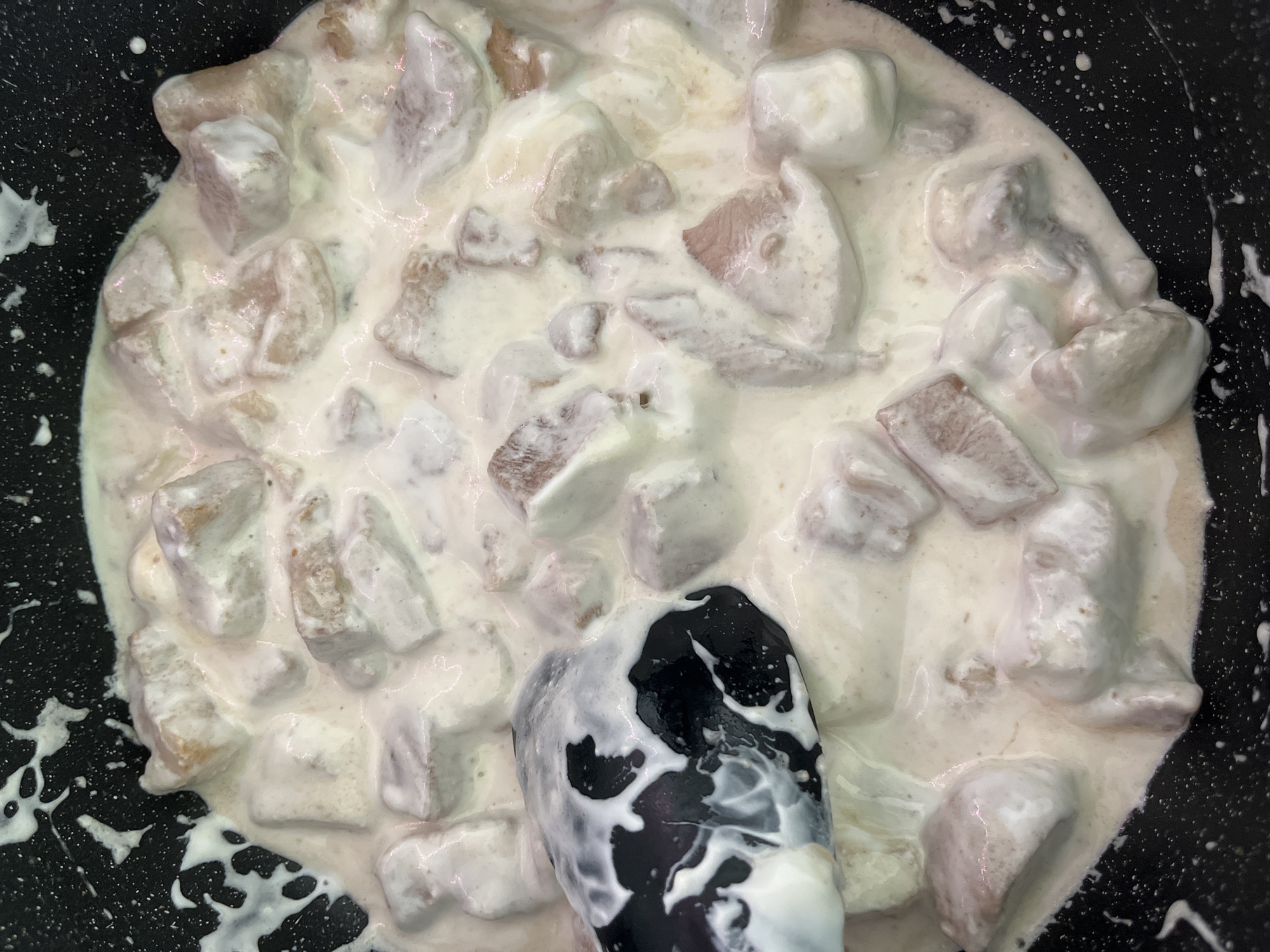 The image shows a close-up, overhead view of food being cooked in a dark, possibly non-stick pan. The food consists of chunks of meat, likely chicken, coated in a creamy white sauce. A black cooking utensil, partially obscured by the sauce, is present in the lower center of the frame. The pan's surface is visible around the edges of the food, revealing a speckled pattern. The scene appears to be indoors, possibly in a kitchen. There is no evidence in the image to connect the scene to Saint Petersburg, Russia, or to any specific individuals.
