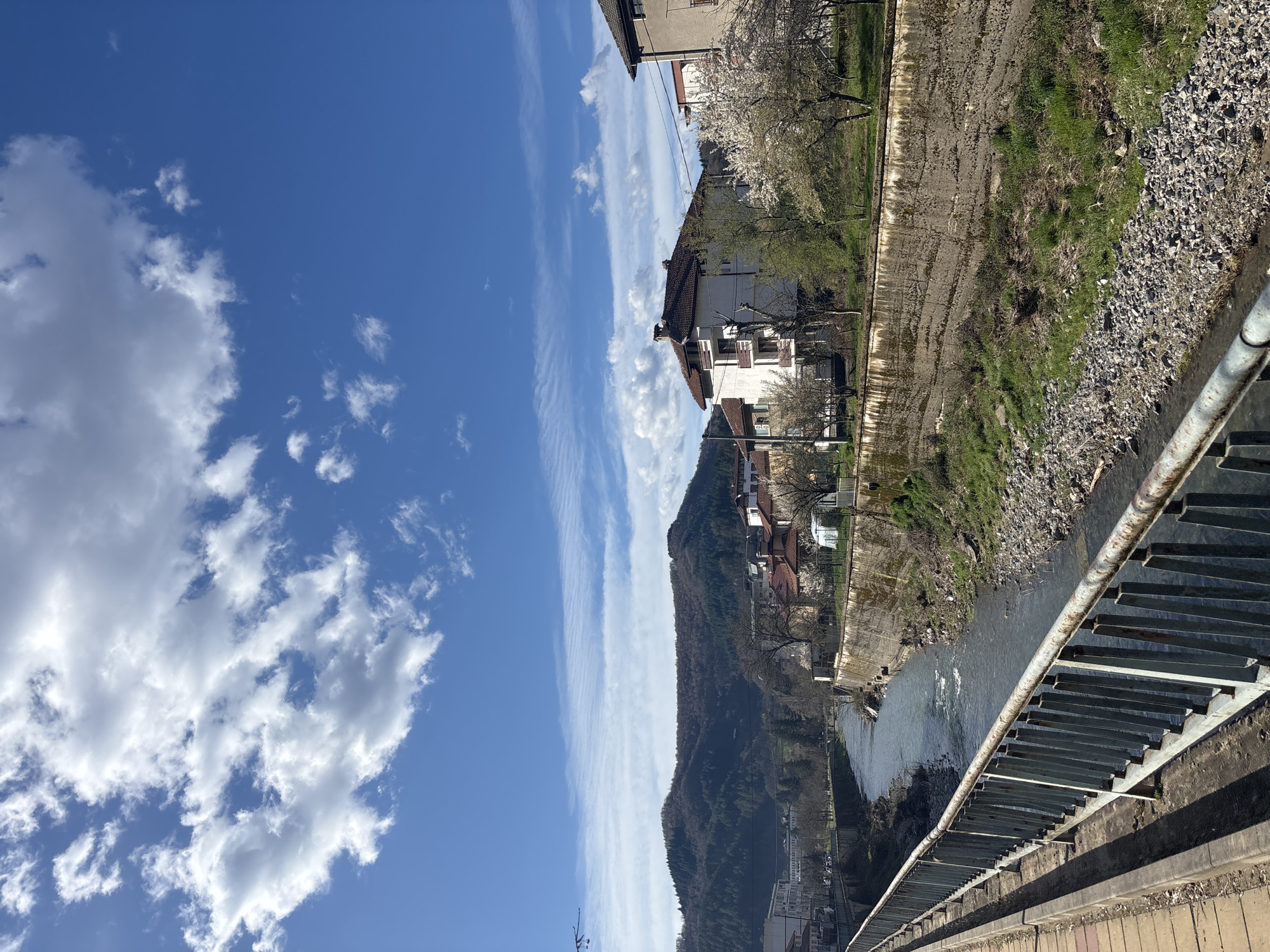 A photograph shows a view of Zlatograd, Bulgaria, oriented on its side. The image foreground features a metal bridge structure running diagonally. A river flows parallel to the bridge, bordered by concrete embankments. Beyond the river, buildings and houses are visible, interspersed with trees and vegetation. A mountain covered in trees forms the backdrop to the town. The sky occupies a significant portion of the frame, exhibiting a clear blue color with scattered clouds.