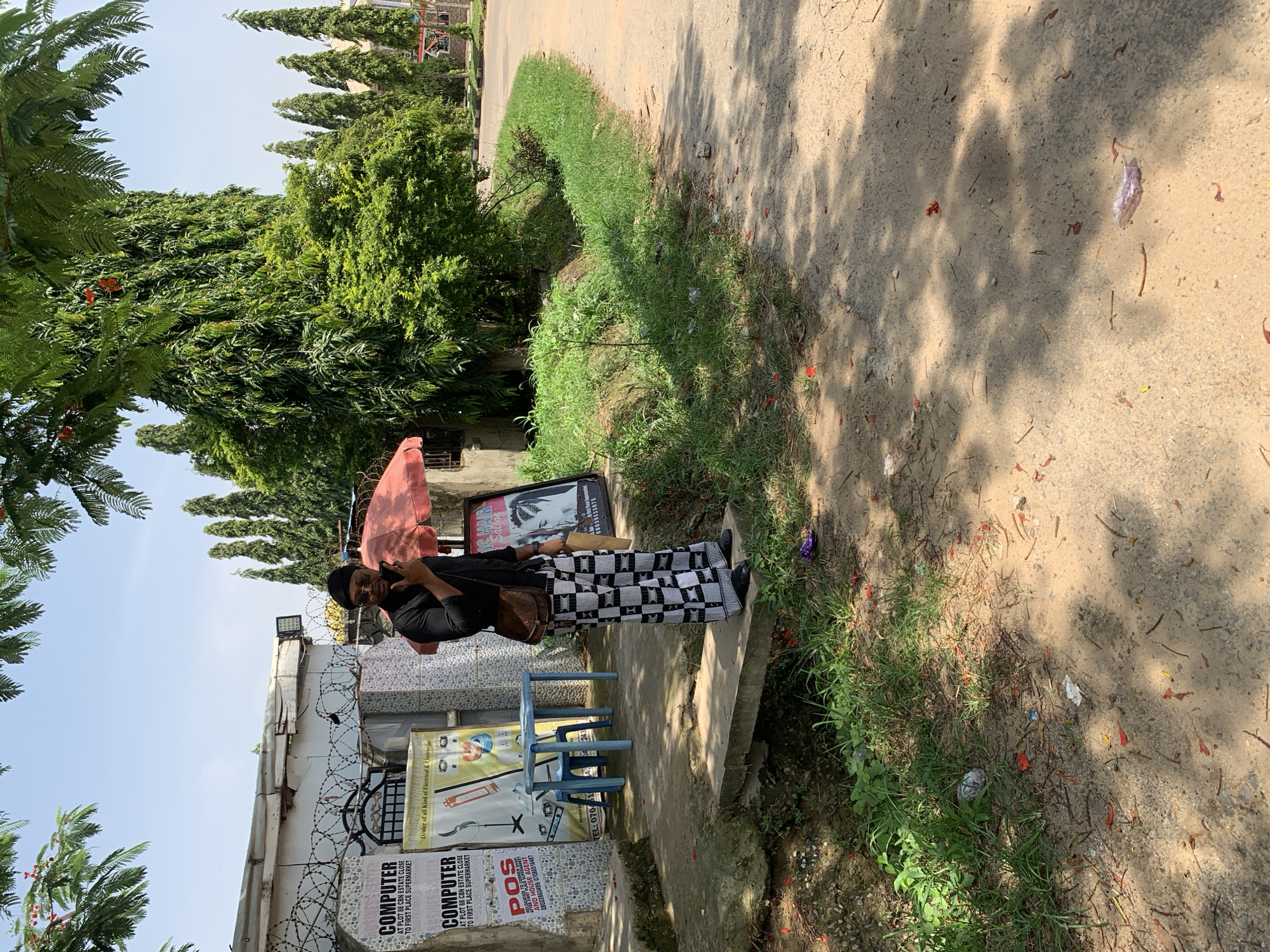 A person wearing a black shirt, black cap, and checkered pants stands on a concrete bridge over a small stream. The person is holding a phone to their ear with their right hand. They are facing the camera and have a brown satchel over their left shoulder. Behind the person is a building with several signs, including one for "COMPUTER" and another for "POS". There are also two small plastic tables and chairs in front of the building. The area is surrounded by lush green trees and vegetation, with a paved road to the right. The scene appears to be an outdoor commercial area in Abuja, Nigeria.