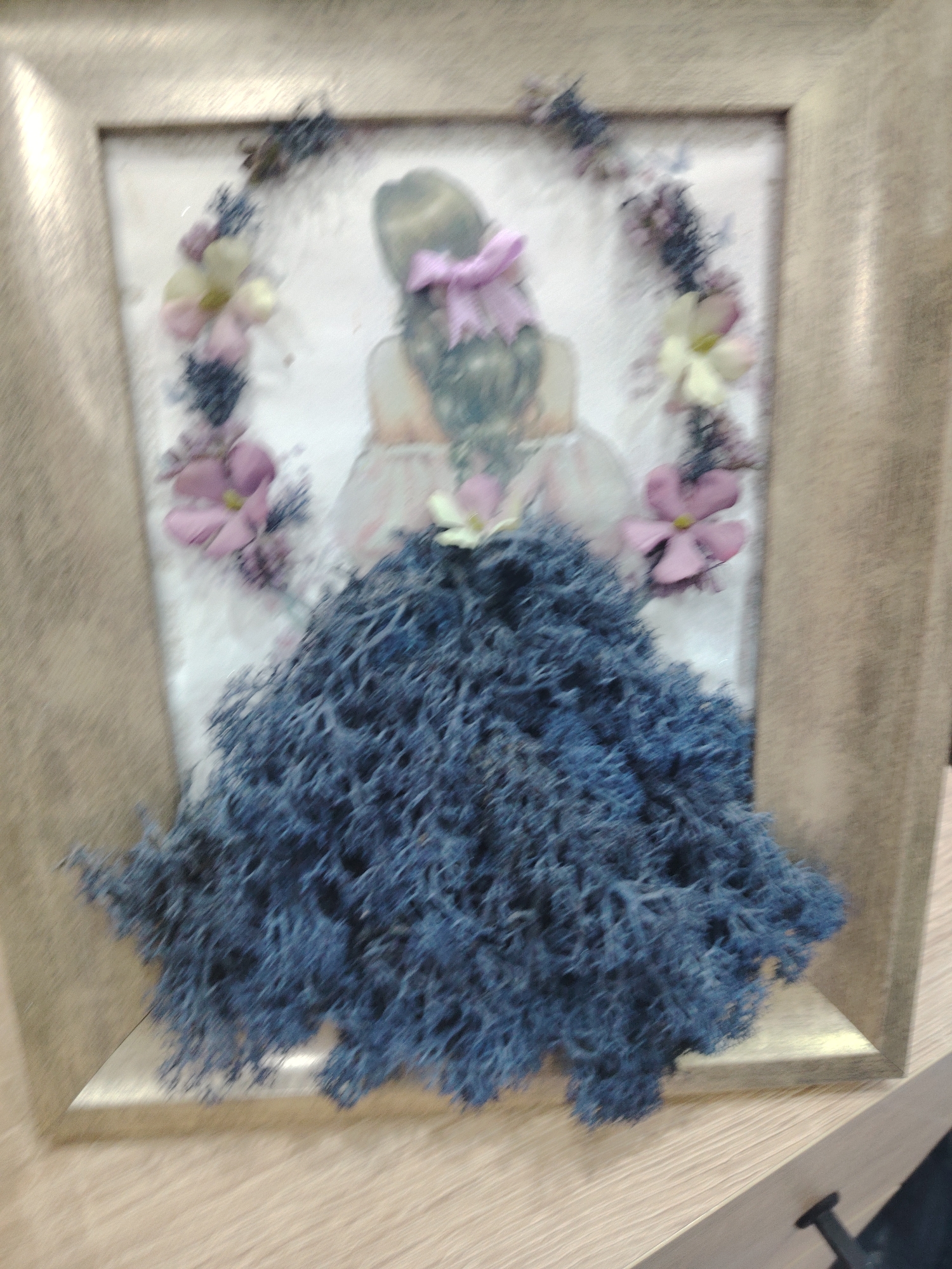 This media file is a framed artwork, likely a decorative piece. The artwork depicts a figure of a girl from the back, created using various materials. The girl's hair is styled with a pink bow. Her dress is represented by a large, voluminous skirt made of a textured, dark blue material that resembles moss or a similar natural fiber. The upper part of the dress appears to be made of fabric or paper, with a light pink color. Around the girl's head is a circular arrangement of small, light yellow and pink flowers and dark, wispy elements, possibly dried flowers or twigs, forming a wreath.
The artwork is housed in a simple, light-colored frame. The background within the frame is a light color, possibly white or off-white, upon which the figure and floral elements are mounted.
The overall impression is a charming and delicate piece of craft art, perhaps intended for home decoration. The focus is on the artistic representation of a girl in a dress, adorned with natural elements. There are no discernible text elements visible in the image. The lighting suggests an indoor setting, and the image quality is slightly blurred, making it difficult to ascertain finer details. The location context provided is Haskovo, Bulgaria, but this is not visually evident from the image itself.