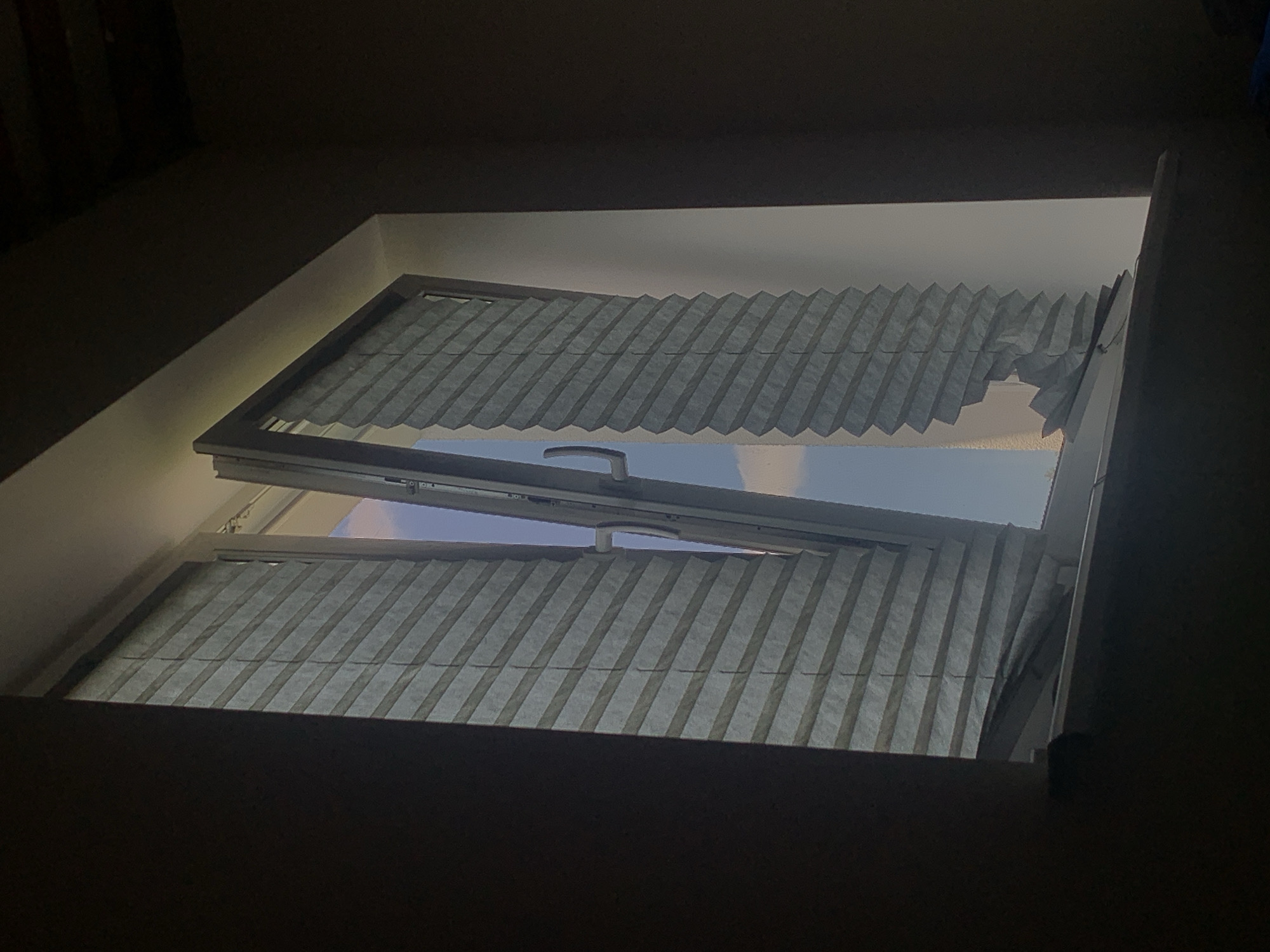 The image shows an interior view of a partially open window with pleated blinds. The window is framed by dark walls, suggesting a room. The blinds on the left are mostly intact and pulled down, while the blinds on the right are pulled up and appear damaged at the bottom, with sections crumpled and detached. Through the open window, a bright sky with some clouds is visible. The scene is lit from the outside, casting shadows within the room. The location is identified as Minsk, Belarus, though no specific landmarks or distinguishing features of the city are visible in the image itself.