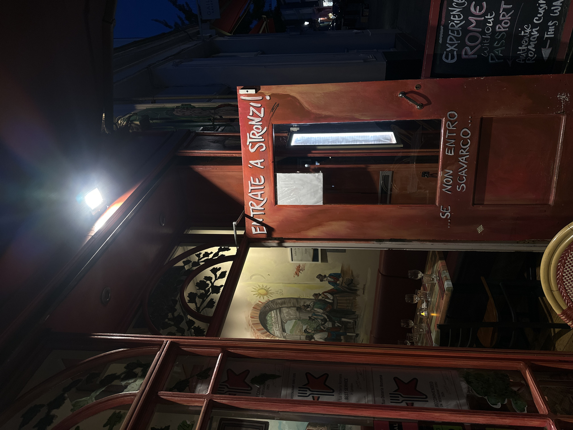 The image shows the entrance of "Tua Osteria Romana," a restaurant in London, United Kingdom. The entrance is decorated with a reddish-brown door frame. On the left side of the frame is the phrase "ENTRATE A STRONZI," painted in white. On the right side, the phrase "SE NON ENTRO SCAVARCO..." is written in white. Above the door frame is a sign that reads "EXPERIENCE ROME without PASSPORT Authentic Roman Cusin." The interior of the restaurant is visible through the glass, revealing a mural of people dining, a table set with glasses, and restaurant seating. A sign advertising the restaurant's awards is displayed in the window.