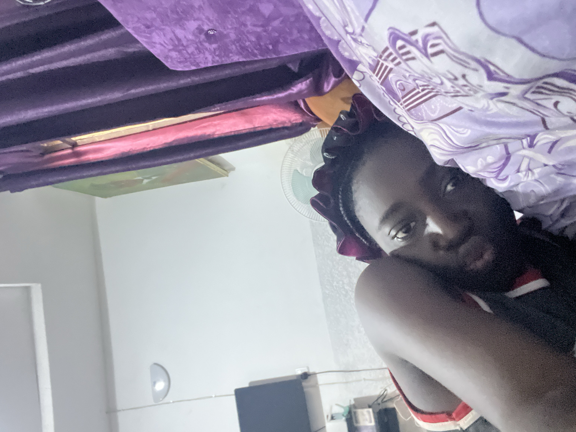 The media file displays an indoor scene, rotated 90 degrees counter-clockwise. In the foreground, a person with dark skin tone is positioned on the right side of the rotated image, looking directly at the camera. Their hair is covered by a dark, possibly purple or burgundy, satin bonnet or scarf with ruffles. They are wearing a dark top with horizontal red and white stripes visible around the shoulder and chest area. Their expression is neutral.
The setting appears to be a bedroom or living space. Directly above the person's head in the rotated image, a light purple patterned blanket or bed sheet is visible. Above this, and occupying a significant portion of the upper left and center of the rotated image, are dark purple curtains, possibly velvet or a similar plush material, arranged in pleats. Below the curtains, a section of lighter, possibly pinkish, fabric or material is seen, along with what appears to be a wooden or light-colored frame structure.
Further in the background, towards the middle right of the rotated image, a white electric fan is partially visible. The majority of the lower left and central background consists of a plain white wall. On this wall, a small, semi-circular light fixture is mounted, and thin cables are strung across it. Below this, a black rectangular object, possibly a speaker or electronic device, rests on a surface. A small framed picture or painting with greenish hues is also discernible on the white wall.
Considering the rotation, the person is likely lying down or reclining. The curtains would be on a wall, possibly covering a window, and the fan and other objects would be on the floor or another wall. The location context is Abuja, Nigeria. There is no visible text in the image.