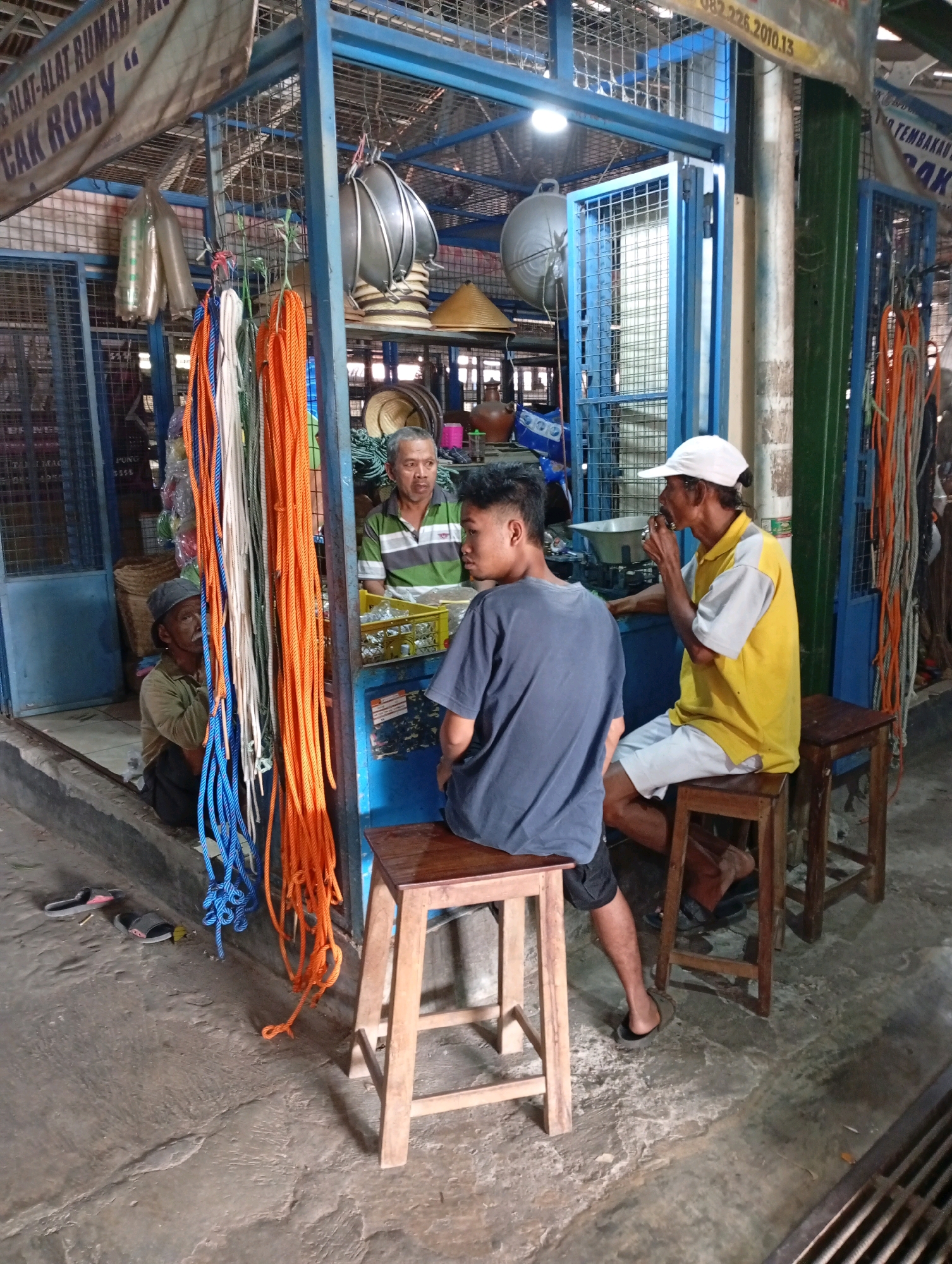 In Playen, Indonesia, a man in a yellow shirt and white cap sits on a wooden stool facing left, holding what appears to be a cigarette. Beside him, a younger man in a dark t-shirt and shorts sits on another stool, facing away from the camera. A third man in a cap and earth-toned shirt sits on the ground to the left, looking up. They are gathered at an outdoor stall with blue metal framing, displaying various items like ropes, baskets, and metal cookware. The ground is concrete, and the overall environment suggests a market or roadside vendor area.