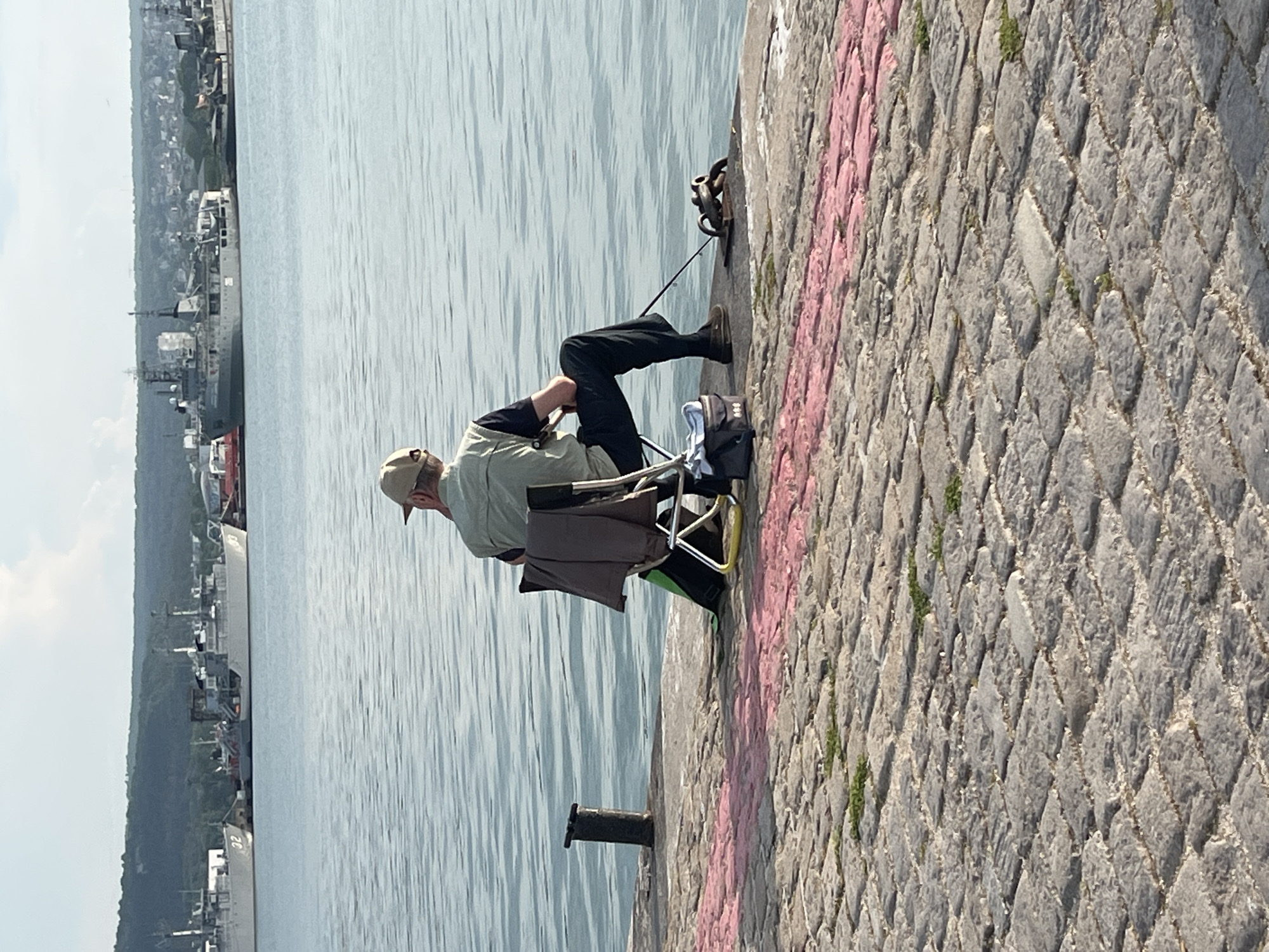 The image portrays a peaceful daytime scene of a person fishing from a stone embankment in Varna, Bulgaria. An older male, clad in a light-colored cap, a light-toned long-sleeved shirt, and dark trousers, is seated comfortably on a portable folding chair. He is actively engaged in fishing, holding a rod that extends towards the calm, gently rippled water, with its tip anchored near large metal rings set into the embankment. A black bag or backpack, with a white cloth, rests beside his chair on the ground.
The embankment is paved with distinctive grey, diamond-patterned cobblestones, with a vibrant pink painted line running parallel to the water's edge. Further along the pier, a black mooring bollard is visible. Across the tranquil water, a distant coastline features a port area with several large ships docked, one bearing the number "32" on its hull. These vessels are backed by lush green hills and scattered buildings, suggesting a coastal or harbor town environment. The bright, soft lighting indicates it is likely late morning or afternoon, under clear or partly cloudy skies, creating a serene and relaxed atmosphere for the fishing activity. The man appears patient and focused on his endeavor.