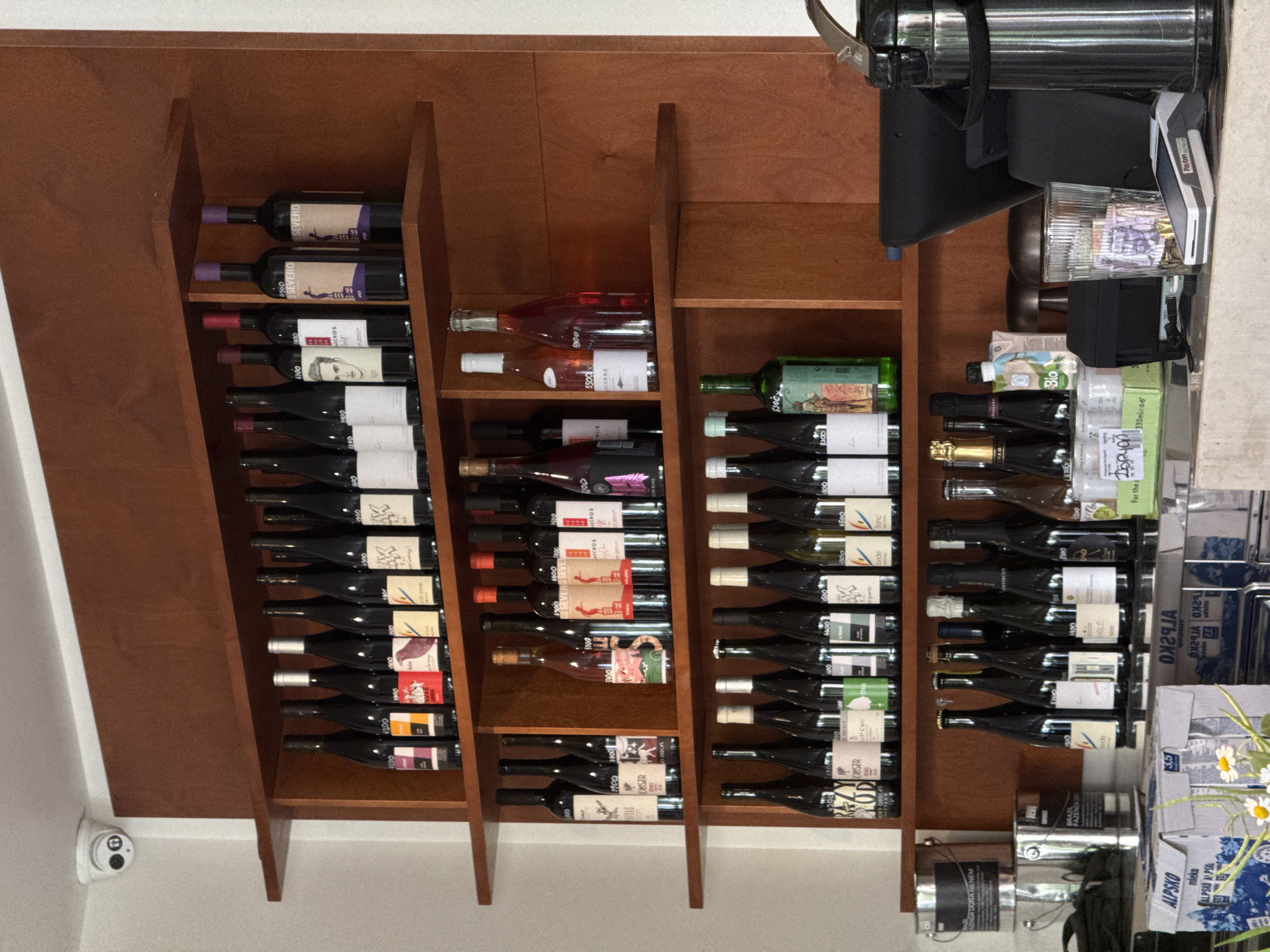 The image shows a shelf in what appears to be a business in Belgrade, Serbia.
The shelf contains multiple bottles of wine, arranged horizontally. A cash register and other supplies are seen adjacent to the shelf. A security camera is mounted on the wall next to the shelf. The brands visible include "OMSER", "SCHOS" and "SEVERO". Also present are milk products.