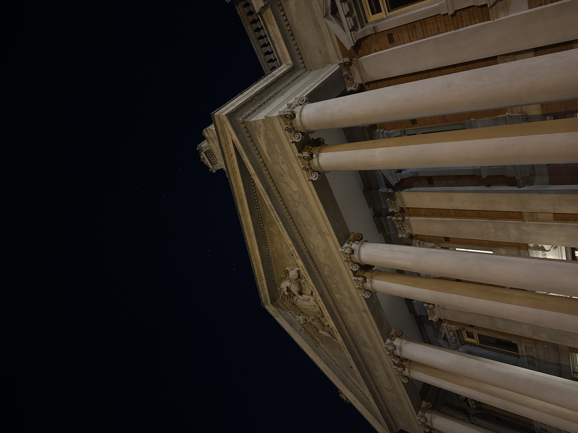 The image displays the illuminated facade of a grand building at night, captured from a low, upward-looking perspective. On the right side, multiple large, light-colored columns are visible, each topped with ornate, scrolled capitals characteristic of classical architecture, such as Corinthian or Composite order. Behind these columns, a red-brick wall is discernible, along with windows set higher up and within the columned structure.
Extending diagonally from the foreground towards the center-left is a prominent architectural element, likely a cornice or entablature. This feature is richly decorated with intricate moldings and a sculpted relief on its underside, hinting at classical mythological or allegorical figures. The lighting strongly highlights the textures and details of the architectural stonework or concrete elements.
The background is a deep, dark black, indicating nighttime. Scattered across the dark sky, particularly in the upper and central-left areas, are faint pinpricks of light, suggesting visible stars in a clear sky. The building itself is brightly lit by artificial sources, creating a stark contrast with the dark night and emphasizing its architectural grandeur. There are no people or discernible activities taking place. No visible text is present in the image. The grand scale and classical design align with architecture found in significant public or historical buildings, potentially within Perth, Australia.