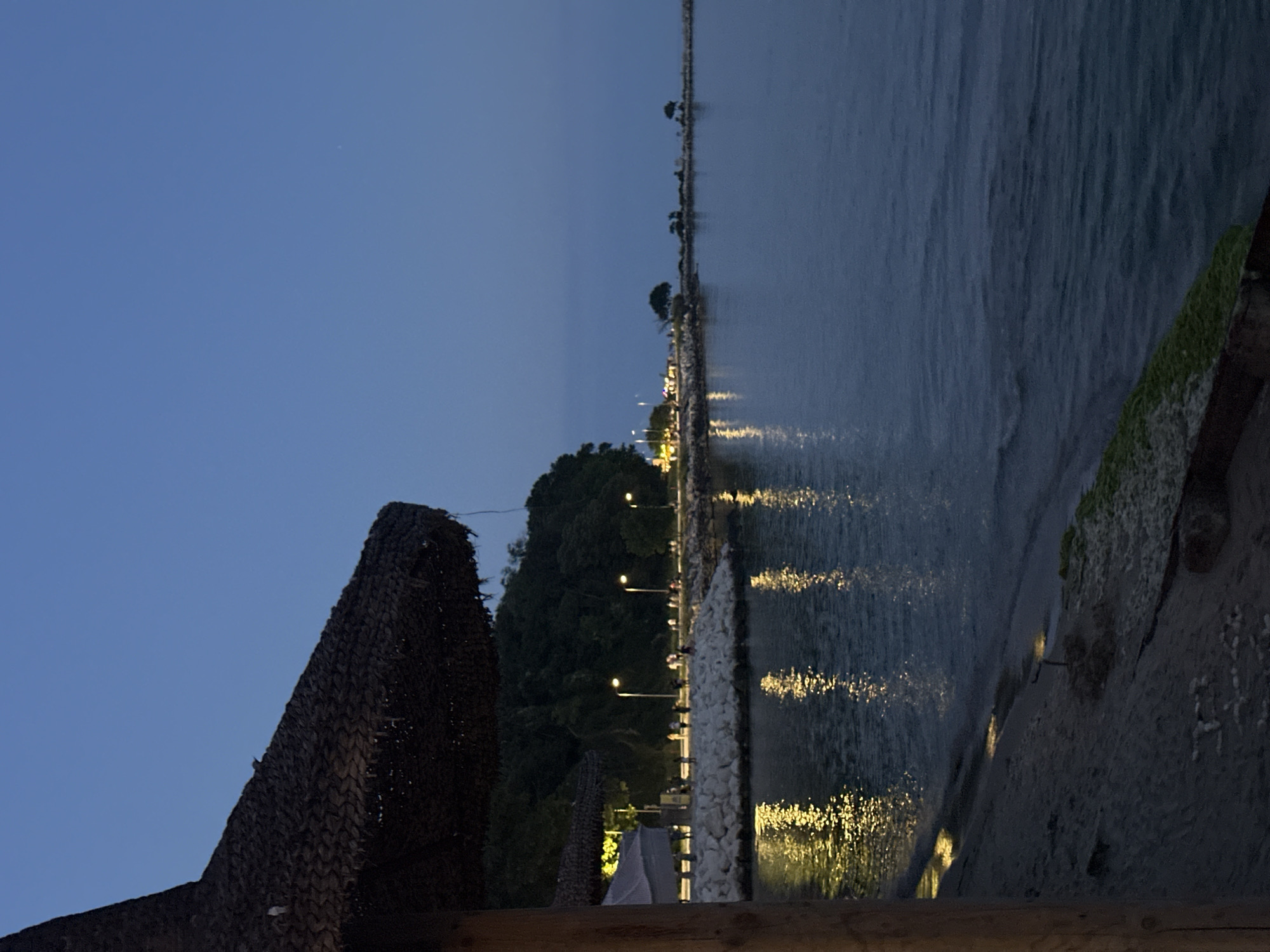 The image depicts a dusky scene at a waterfront promenade. In the foreground, a large, thatched umbrella dominates the left side of the frame, casting a shadow over a sandy beach and the water's edge. The water is calm, reflecting the warm glow of streetlights lining the promenade. A stone breakwater extends into the sea, with a line of trees and vegetation visible on the land behind it. Several people are strolling along the promenade, illuminated by the streetlights, suggesting a leisurely evening atmosphere. The sky is a deep, clear blue, transitioning to darkness as night approaches. The overall impression is one of a serene evening by the sea, with a hint of activity from the people enjoying the waterfront.