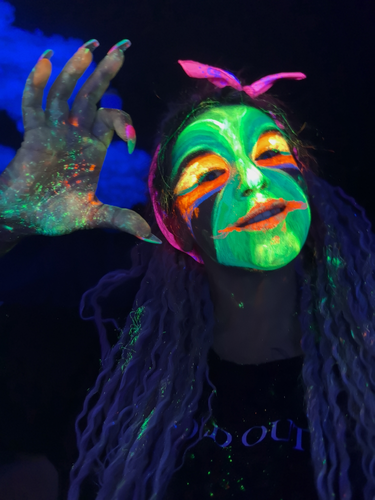 The image depicts a person with elaborate neon-colored face paint in a dark environment. The face paint features bright green, orange, blue, and red colors. The person is wearing a pink headband with bunny ears. They have long blue braids. The person's left hand is raised, displaying the same neon colors. The person is wearing a black shirt with the words "OUT" in neon blue. The location is not discernible from the image.