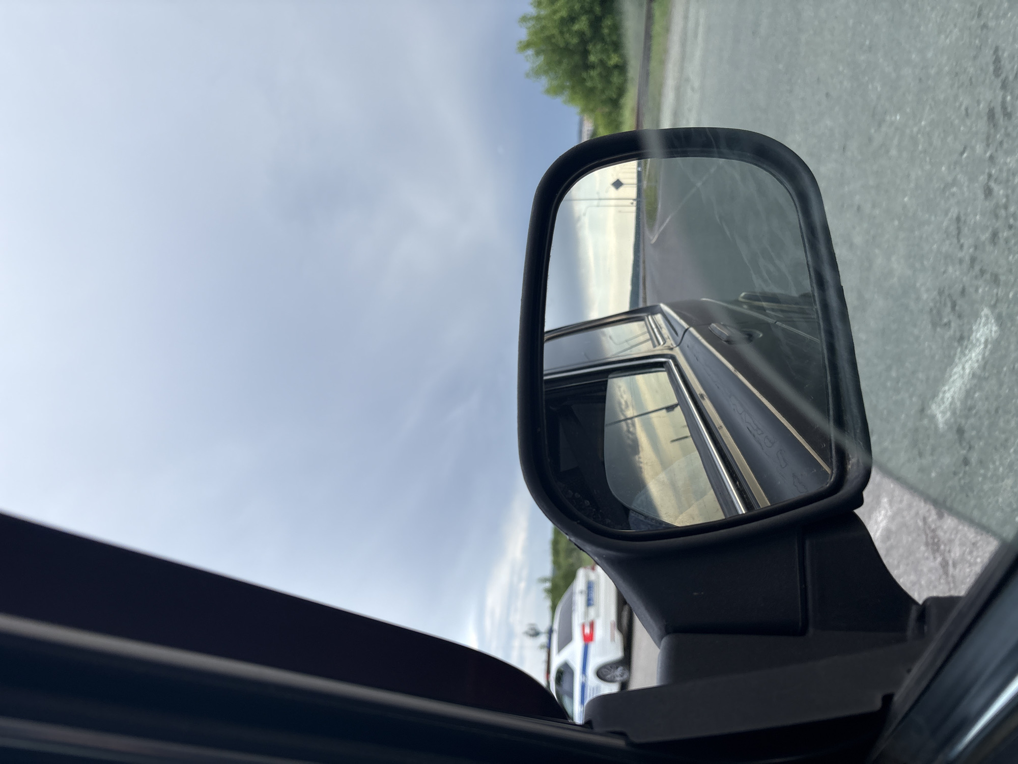 The image, taken from inside a vehicle, shows a car's side mirror reflecting a road and the car's side. A white vehicle with blue striping and a red taillight is visible behind the car, possibly a police vehicle. Outside the window and in the mirror, there is a cloudy sky and some greenery. The location is described as Chaikovsky, Russia. The surrounding area appears to be a paved parking area.