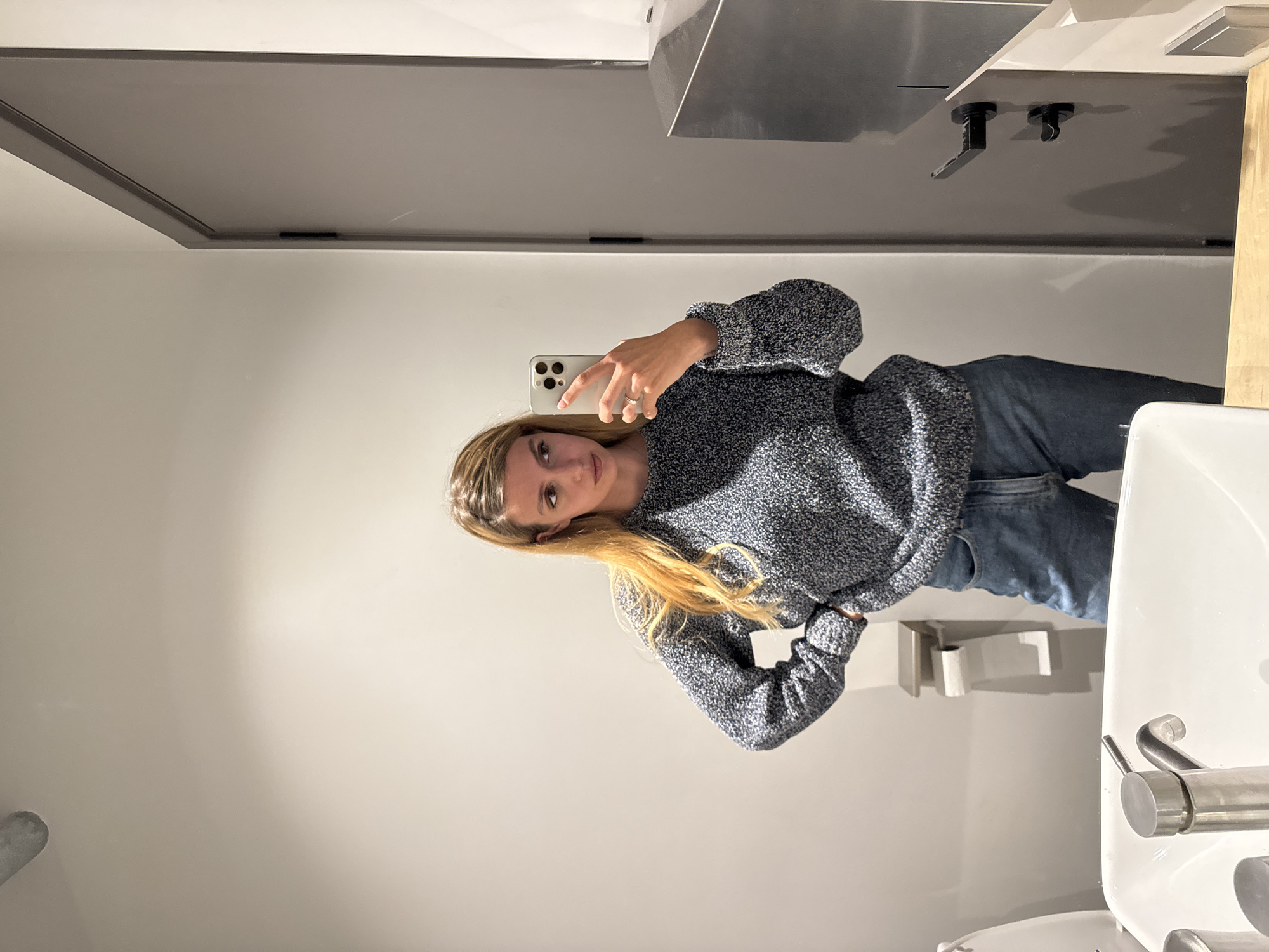 A woman takes a selfie in a bathroom mirror. She is wearing a blue and gray sweater and blue jeans. The bathroom has a gray door, a stainless steel sink, a white sink, and a stainless steel toilet paper holder. The location is in the city of Unknown, Unknown.