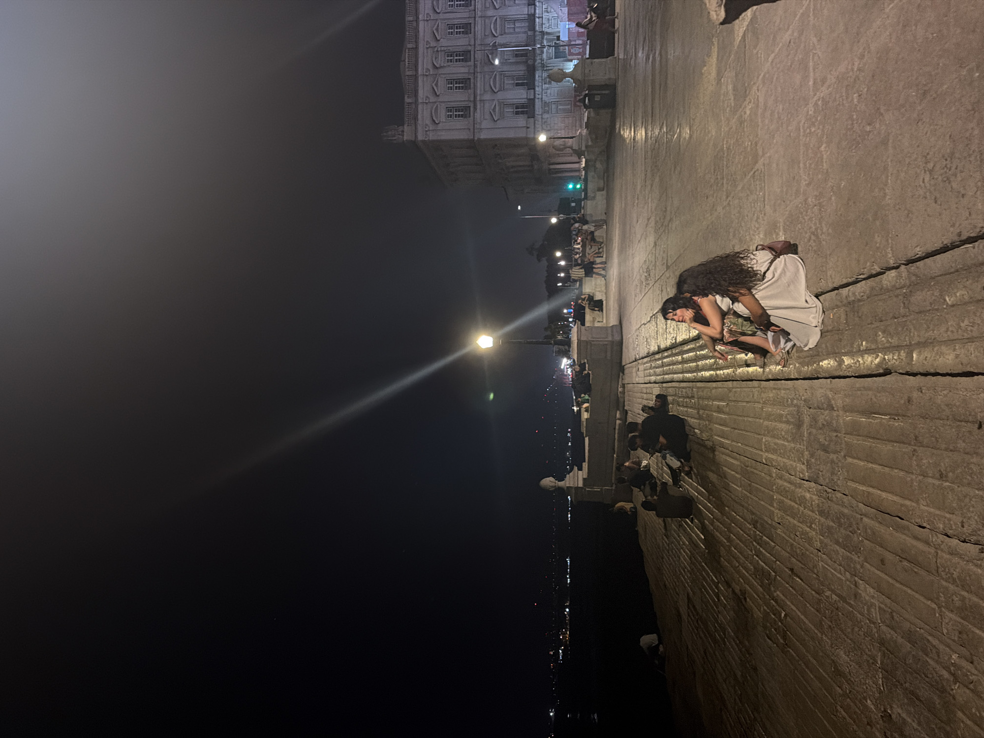 The photograph depicts a nighttime scene along a waterfront promenade in Lisbon, Portugal, likely near the Praça do Comércio or Cais das Colunas given the architectural style and proximity to the Tagus River. Two women, one with dark curly hair and wearing a light-colored dress, and another with dark hair and wearing a patterned dress, are seated together on the stone pavement in the foreground. The woman in the light dress is leaning back, while the other appears to be looking out towards the water. A brown bag is visible near them.
Further in the background, several other individuals are scattered across the stone walkway, some sitting in groups and others walking. To the left, a dark body of water is visible, with distant city lights reflecting on its surface, suggesting a shoreline across the river. A large, ornate building with multiple windows and classical architectural features stands on the right side of the frame, illuminated by streetlights. Street lamps with glowing bulbs are also present, casting long beams of light across the scene. The ground is paved with large, rectangular stones, showing signs of wear. The overall atmosphere is serene, with a sense of urban nighttime ambiance.