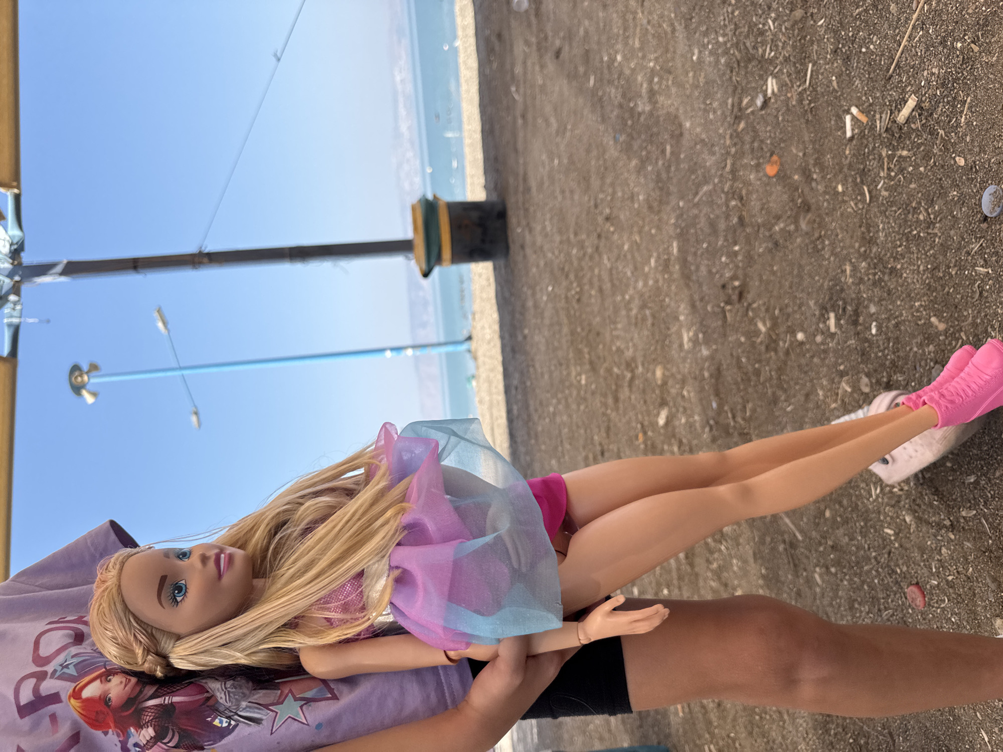 This media file depicts a Barbie doll lying on a person's lap, outdoors. The setting appears to be a beach or a promenade near a body of water, with a clear blue sky and a concrete or gravel ground scattered with small debris like cigarette butts. The doll is wearing a pink swimsuit and a sheer, multi-colored pink and blue dress. The person holding the doll is wearing a purple t-shirt with graphics of animated characters and black shorts. The time of day is likely daytime, given the bright sunlight and blue sky. The overall impression is a casual, playful moment, possibly at a beach resort or a park. There is no visible text of significance in the image.