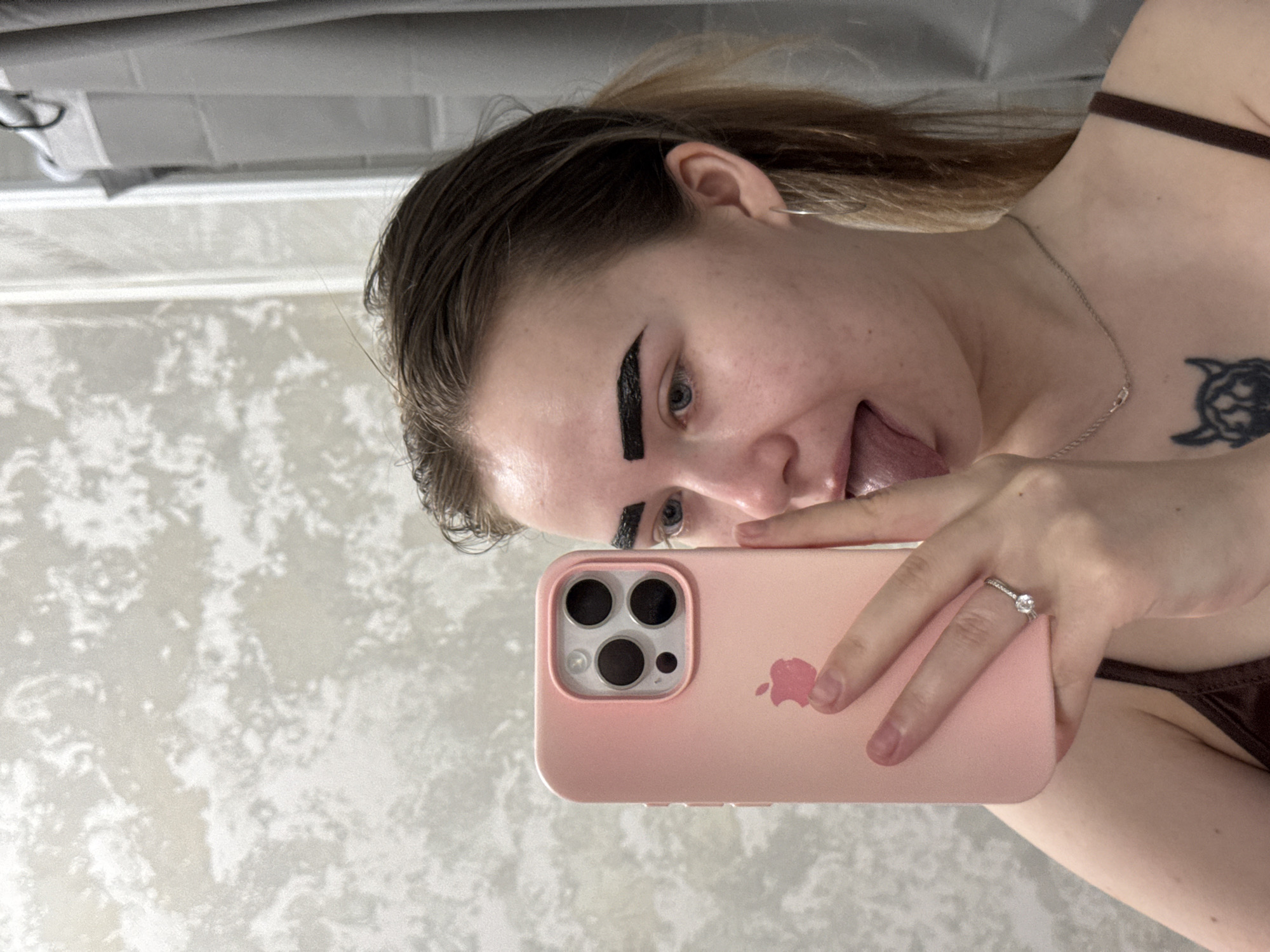 A young woman is taking a selfie in what appears to be a bathroom. She is holding a pink iPhone in her right hand, and a ring is visible on her ring finger. She is sticking her tongue out. She has dark substance applied to her eyebrows. She has a tattoo on her chest. The background includes a white and gray patterned wall and what appears to be a shower curtain rod. There is no evidence to confirm that the location is Chaikovsky, Russia.
