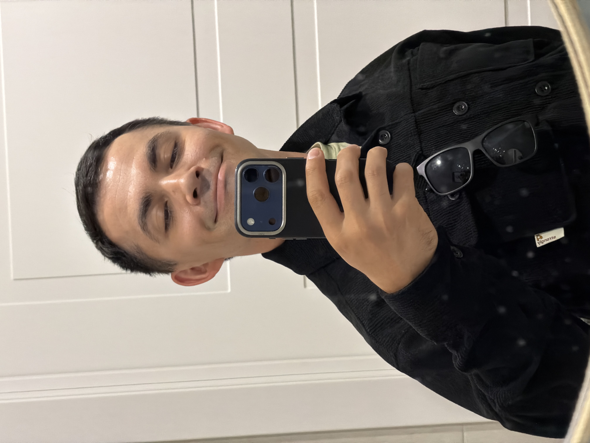 A person is taking a selfie in a bathroom with a smartphone, likely an iPhone, visible in the reflection. The individual is wearing a dark, button-down corduroy shirt, and a pair of sunglasses is tucked into the shirt. The background shows a white paneled door. No information suggests this image was taken in Izhevsk, Russia.
