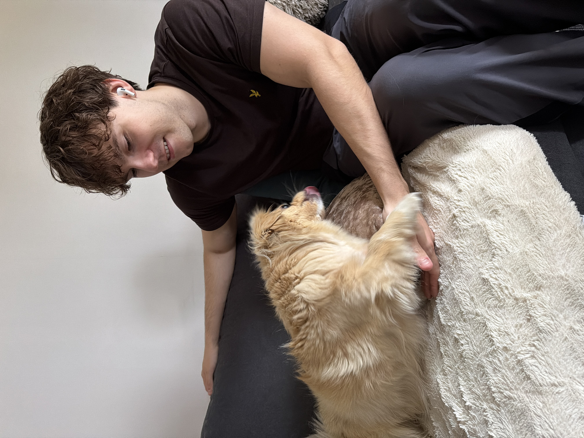 A young man with curly brown hair and white earbuds is sitting on a dark grey couch, looking down at a light brown, fluffy dog that is partially on his lap. The man is wearing a dark brown t-shirt and dark grey pants. The dog is licking its tongue and its paw is resting on the man's hand. A fluffy white blanket is spread across the couch in front of the dog. The background is a plain white wall. The scene appears to be an indoor living room. There is no indication of the city of Harstad, Norway.