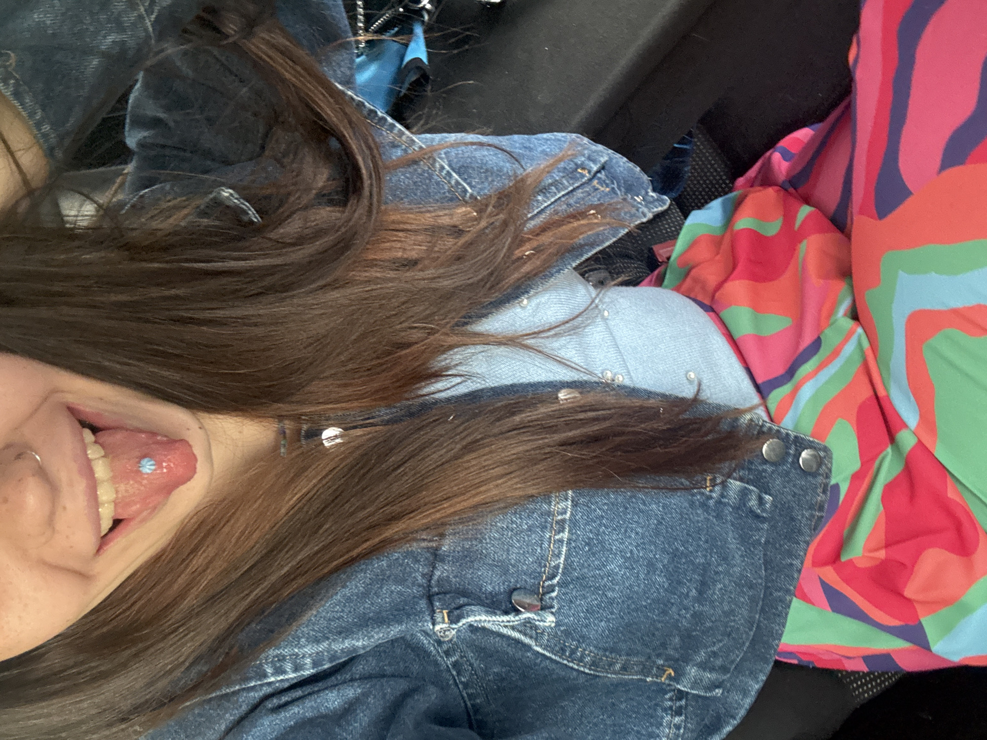 A close-up, vertically oriented image captures a person's lower face, shoulder, and part of their torso, possibly taken as a selfie. The person's mouth is open, and their tongue is extended, revealing a light blue, flower-shaped tongue piercing. A small silver ring is visible on the side of their nose. Long, brown hair cascades over their shoulder and arm.
The person is wearing a dark blue denim jacket with silver buttons, partially open to show a lighter blue garment underneath, which also has small silver studs or embellishments. To the right of the person, a vibrant, abstractly patterned fabric is visible, featuring swirls of pink, red, green, light blue, and purple. The background behind the person is dark and indistinct, suggesting an interior setting, possibly a car seat or dark furniture. The overall mood conveyed is casual and playful. There is no visible text in the image. The location context provided is Sofia, Bulgaria.
