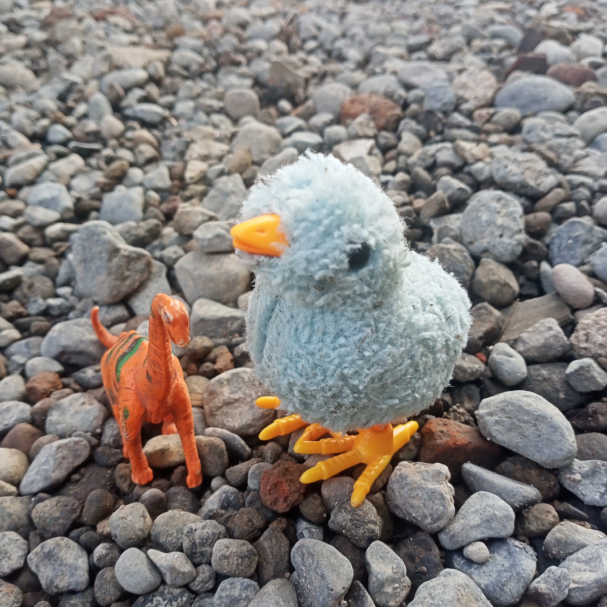 An orange toy dinosaur with green stripes stands to the left of a light blue plush toy chick with orange feet and beak. Both toys are positioned on a surface covered with small, gray and brown stones. The background consists of more stones, out of focus. The scene appears to be outdoors.