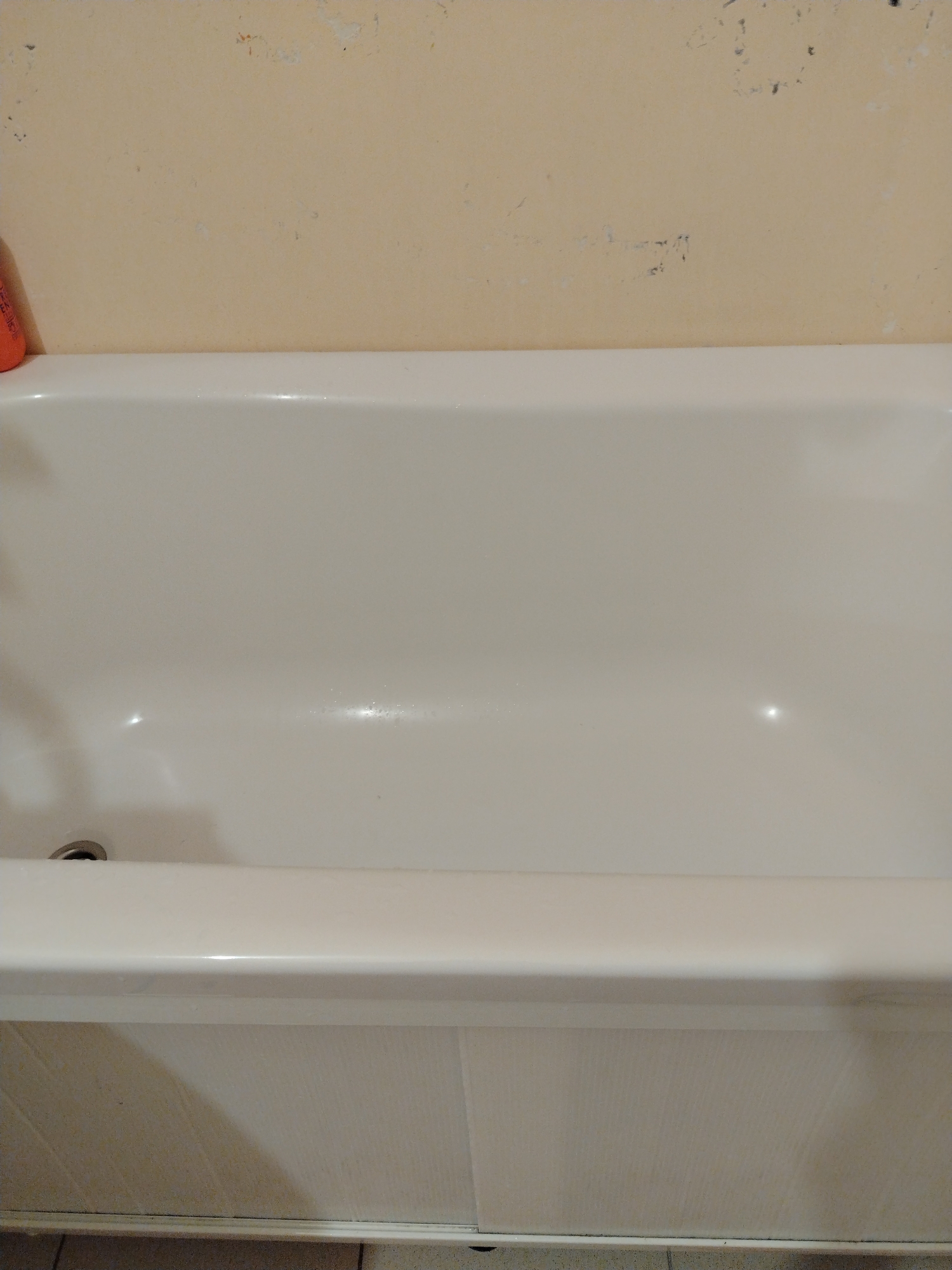 The scene depicts an empty, white bathtub and the adjacent wall, located in the city of Смалявічы, Belarus. The lower two-thirds of the image display the interior of the bathtub, which has a glossy, off-white surface with minor reflections and what appear to be small water droplets. A dark, circular drain is visible in the lower-left corner of the tub's basin. The upper third of the image shows a light beige-colored wall above the bathtub, exhibiting signs of wear such as discolored patches, peeling paint, and faint dark marks. A small portion of an orange object, resembling a container, is partially visible on the far-left edge, resting on the bathtub rim against the wall. The lower edge of the bathtub's exterior panel, a light-colored, possibly tiled or paneled surface, is visible at the very bottom of the frame, with a sliver of darker flooring directly beneath it. No individuals are present.
