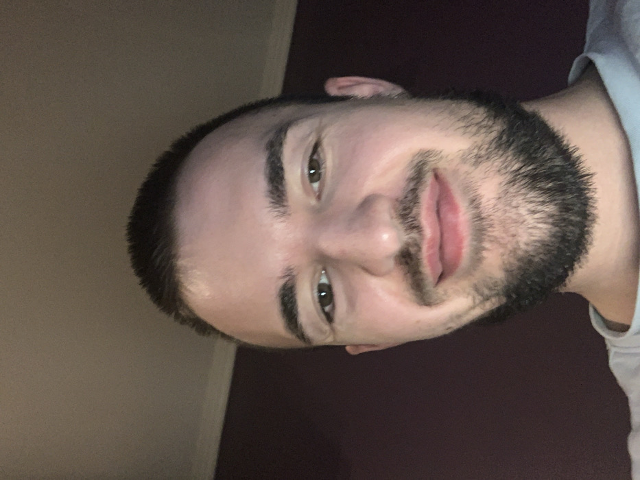 A male individual is depicted in a close-up, head-and-shoulders shot, presumed to be a selfie. The subject has short, dark hair and a neatly trimmed dark beard covering the chin and jawline. He possesses dark eyebrows and brown eyes, directed straight at the viewer. A faint smile is present on his closed lips. He is wearing a light-colored crew-neck shirt, visible at the bottom of the frame. The background indicates an indoor setting, with an upper section displaying a light beige or off-white ceiling or upper wall, featuring a visible molding strip. The lower background, behind the subject's left side, consists of a solid, dark purple or burgundy wall. Illumination appears to be artificial, casting some highlights on the forehead. The scene is understood to be located in Sofia, Bulgaria, although no specific city landmarks or identifiers are visible within the frame.
