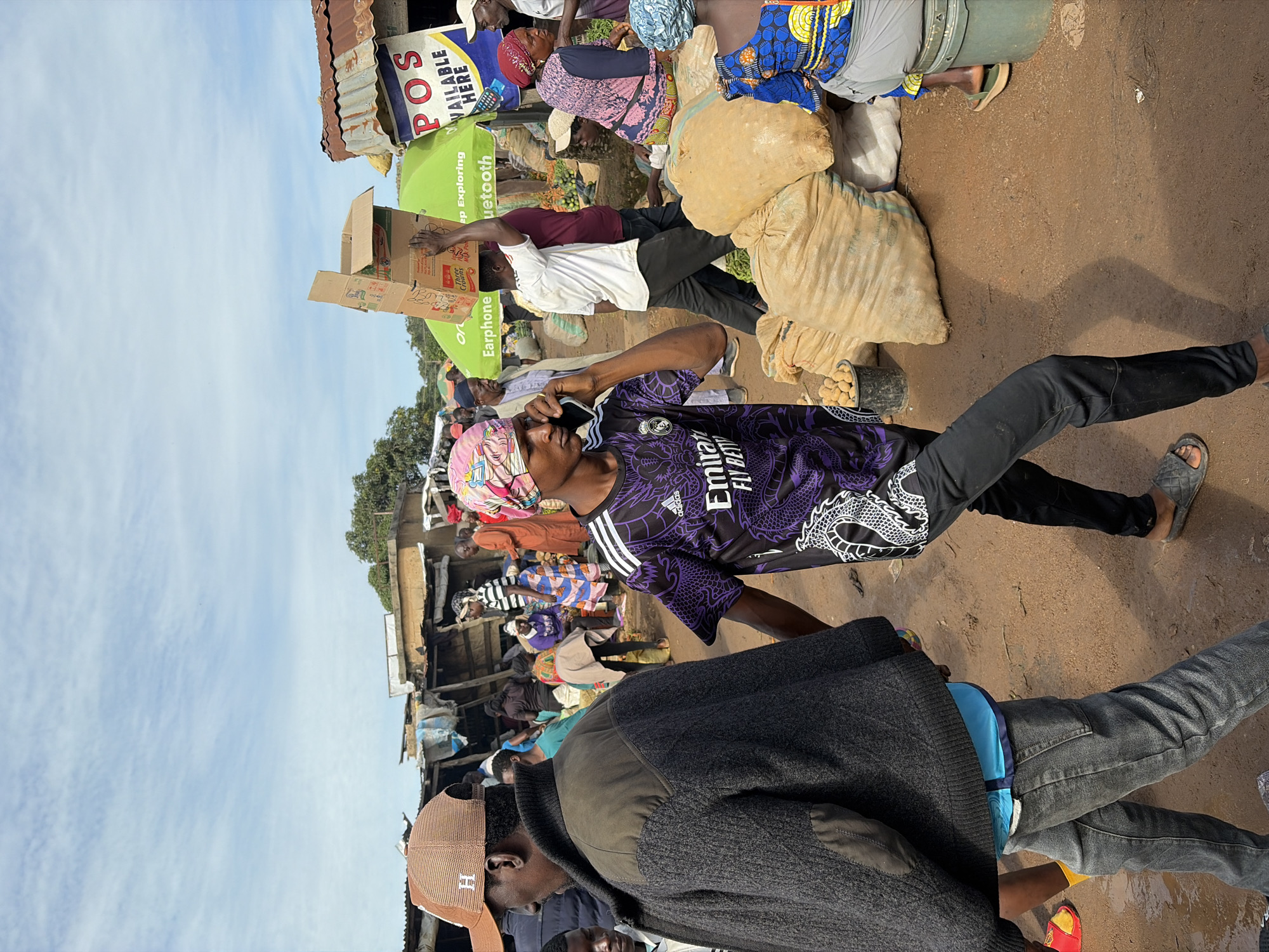 The image depicts a busy marketplace scene in Gura Top, Nigeria. Several individuals are present, engaged in various activities. In the foreground, a man wearing a dark jacket and jeans walks to the left, while another man in a purple t-shirt and black pants talks on a mobile phone. Behind them, a man carries a cardboard box on his head. Various stalls and structures, some with corrugated metal roofs, line the background, indicating a typical market environment. Bags of produce, likely potatoes, are stacked on the ground, and a green umbrella with visible text is set up. The ground is dusty and uneven, with some areas appearing wet. The sky is partly cloudy.