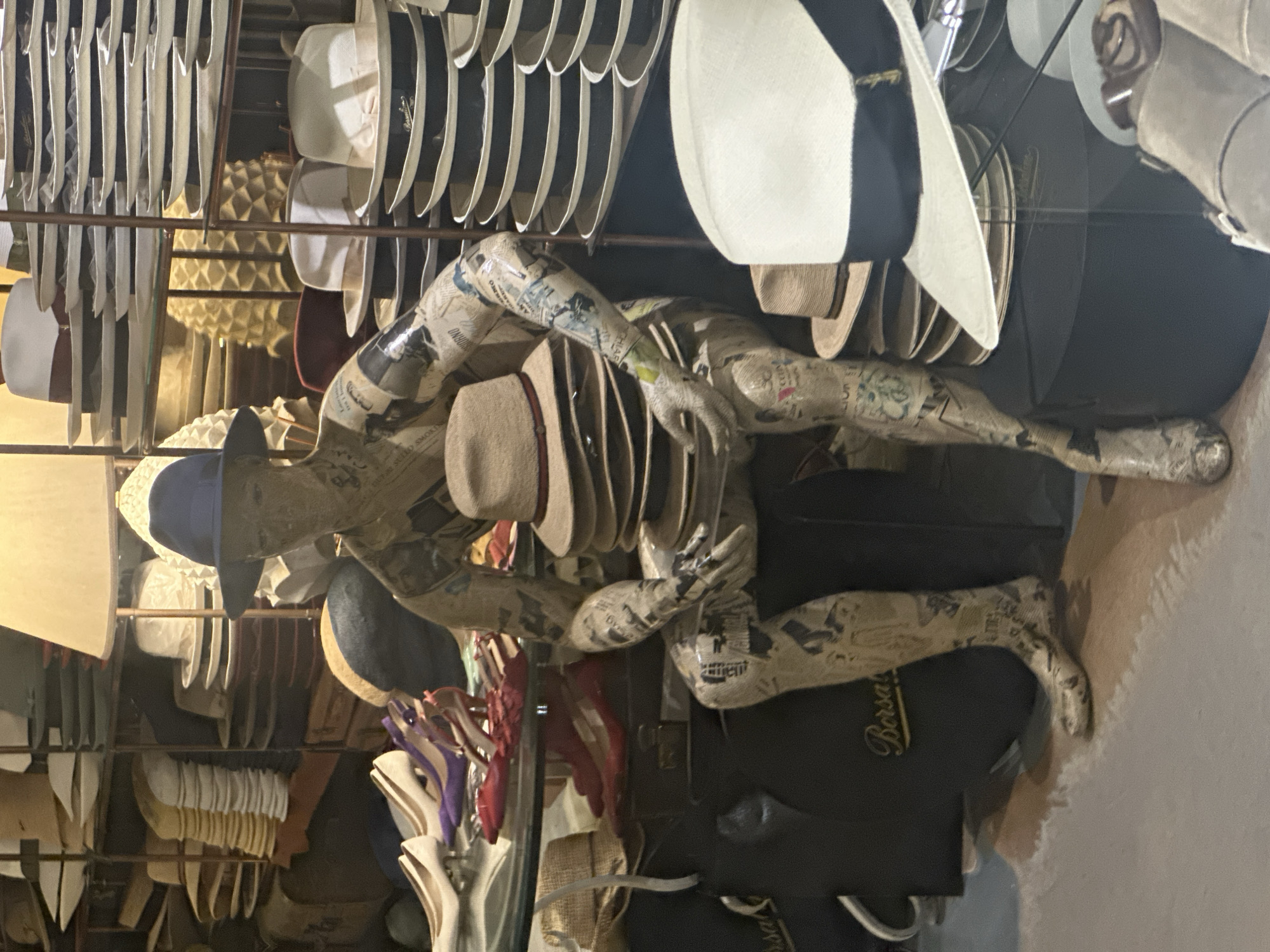This image, viewed upright, depicts an interior scene from a stylish hat shop, likely in Taormina, Italy. The setting is filled with an extensive display of various hats and a unique, artistic mannequin serving as a central focal point.
Dominating the foreground is a life-sized mannequin, distinctive for its surface covering of newspaper print, creating a textured, collage-like appearance. It is posed in a relaxed, seated or reclining position, wearing a light blue baseball cap. Another light brown fedora-style hat with a dark band rests casually on its arm.
The background and surrounding areas are filled with a wide array of hats. Numerous light-colored hats, resembling Panama hats or fedoras, are neatly organized on multiple shelves and racks, suggesting a well-curated retail display. These are interspersed with hats in various other colors, including white, black, and touches of blue and red, some displayed on individual stands.
A notable detail in the lower right corner is a dark bag or box bearing the prominent brand name "Borsalino" in elegant gold script. This identifies a renowned Italian hat manufacturer, strongly aligning with the provided location context of Italy. The shop is illuminated by artificial lighting, creating a warm ambiance and highlighting the craftsmanship of the headwear on display. The scene evokes the atmosphere of a specialized boutique dedicated to high-quality hats.