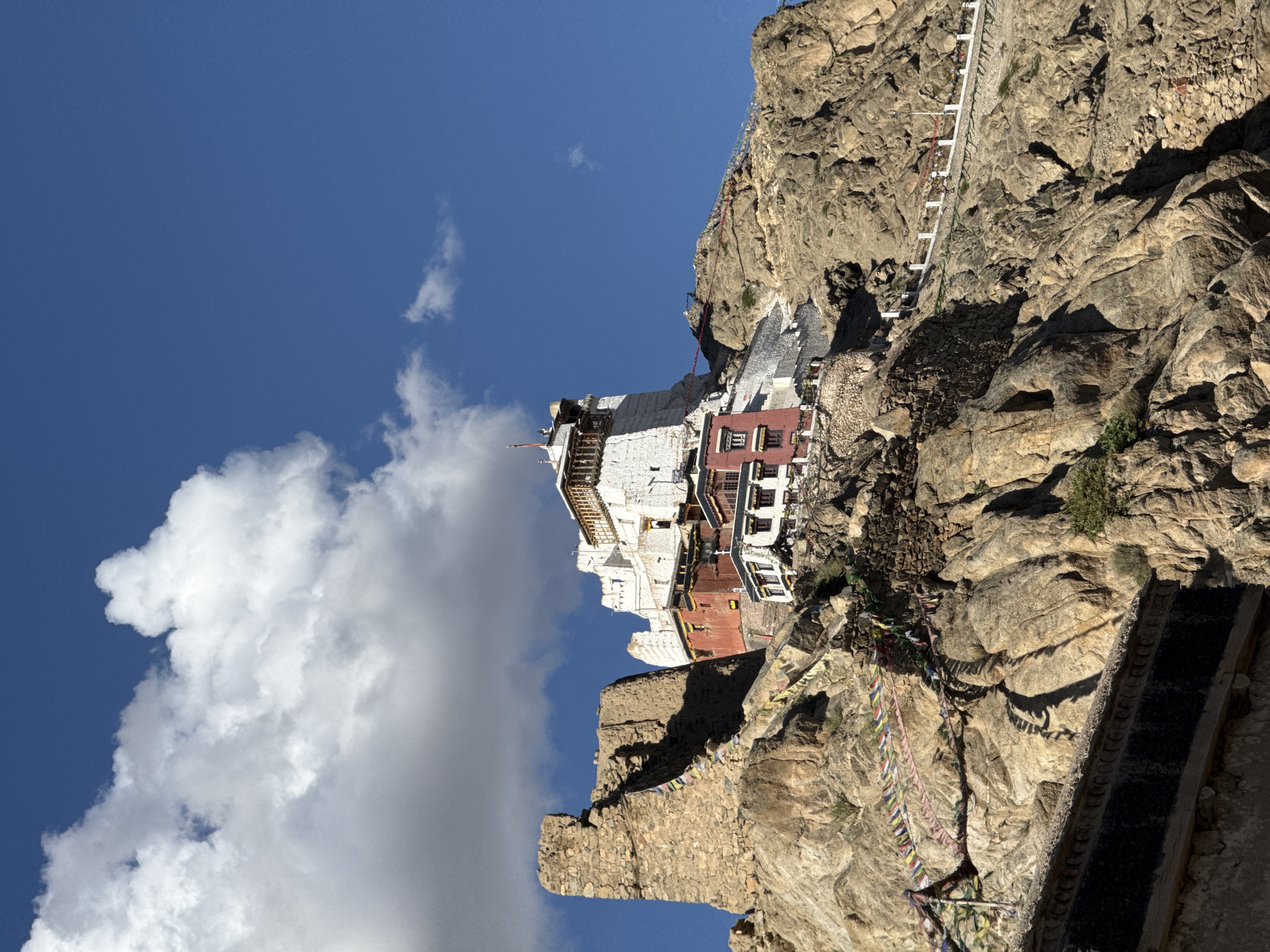 This image depicts the Tsemo Monastery complex, also known as Tsemo Fort, perched atop a rocky hill in the city of Leh, India. The monastery, a prominent landmark, features white-washed buildings with red accents, topped with traditional tiered roofs and prayer flags. The architecture is characteristic of Tibetan Buddhist monasteries. The monastery is situated on a rugged, arid hillside composed of layered rocks and stones. Prayer flags, multi-colored and strung across the hillside, are visible throughout the scene, fluttering in the wind. The sky above is a vibrant blue, with scattered white clouds, suggesting clear weather. The lower part of the image shows the base of the hill and some of the surrounding rock formations, with a dark, textured wall in the foreground. The scene is captured from a low angle, emphasizing the imposing height and scale of the monastery and its mountainous setting. There are no people visible in the frame.