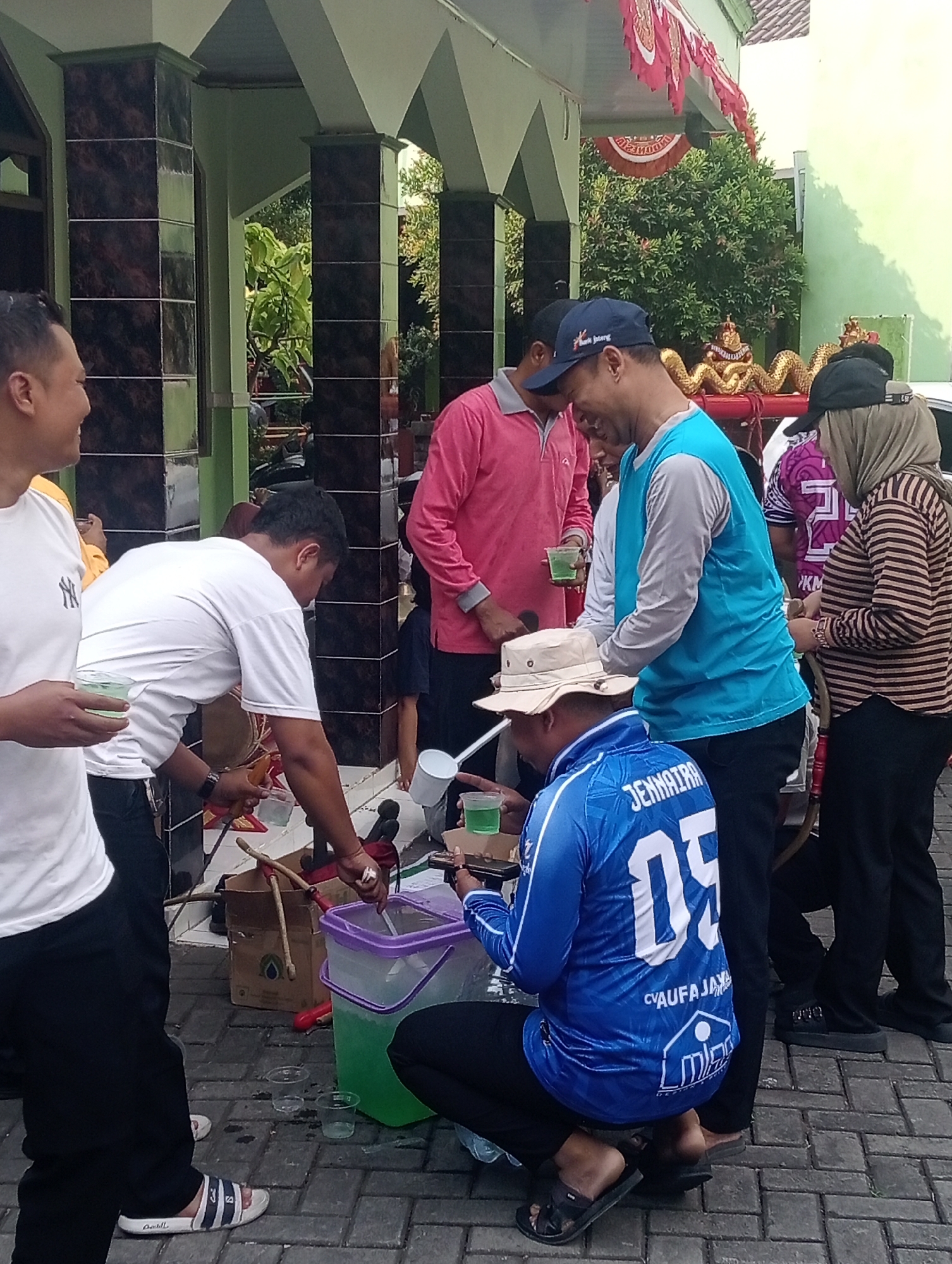 In Demak, Indonesia, a group of people are gathered outside, seemingly at an event or public space. A man in a bright blue vest over a light grey long-sleeved shirt is pouring a green liquid from a white pitcher into a cup held by a person to his left. Another man, wearing a pink long-sleeved shirt, stands nearby, holding a clear cup with green liquid. A man in a white t-shirt is on the left of the frame, holding a clear cup with green liquid, and looking to his right. A person wearing a light-colored hat and a blue long-sleeved shirt with white lettering is squatting in the foreground, also with a cup of green liquid. To the right, a woman in a hijab and a striped long-sleeved shirt is visible. In the background, there are pillars with dark tiled accents and lush green trees. A red and white banner is draped above. The ground is paved with brick-like material. Several plastic containers and cups are present, suggesting a distribution or serving of beverages.