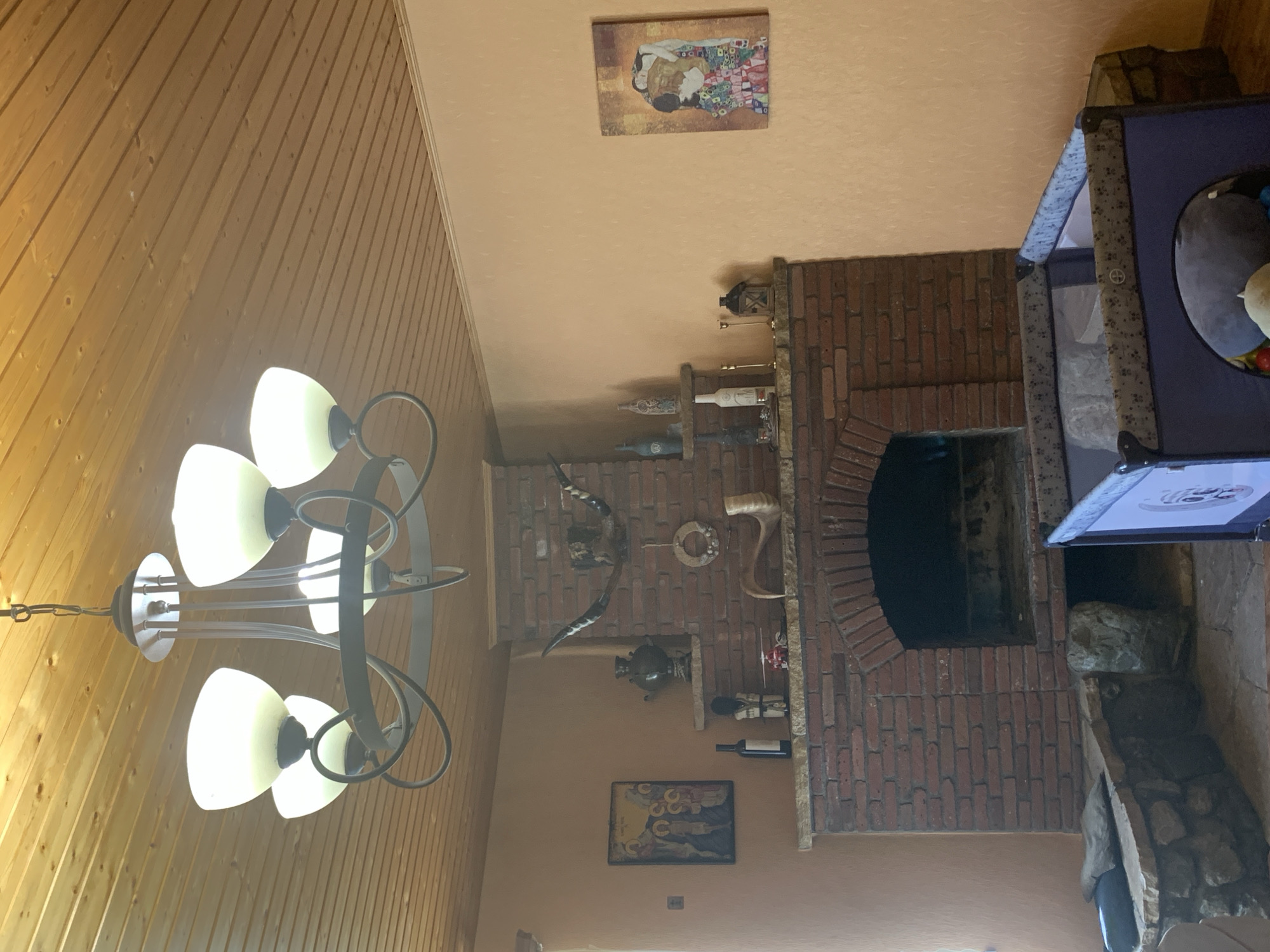 This image depicts the interior of a room with a brick fireplace and a wooden ceiling. A chandelier hangs from the ceiling. On the mantelpiece above the fireplace are various decorative items, including a samovar, animal horns, and a small toy helicopter. To the left of the fireplace, there is a framed religious icon and a bottle of wine. To the right of the fireplace, on the wall, is a framed print of "The Kiss" by Gustav Klimt. In the foreground, to the right, is a portable crib, and to the left, there is a stone bench. The overall aesthetic suggests a rustic or traditional decor.