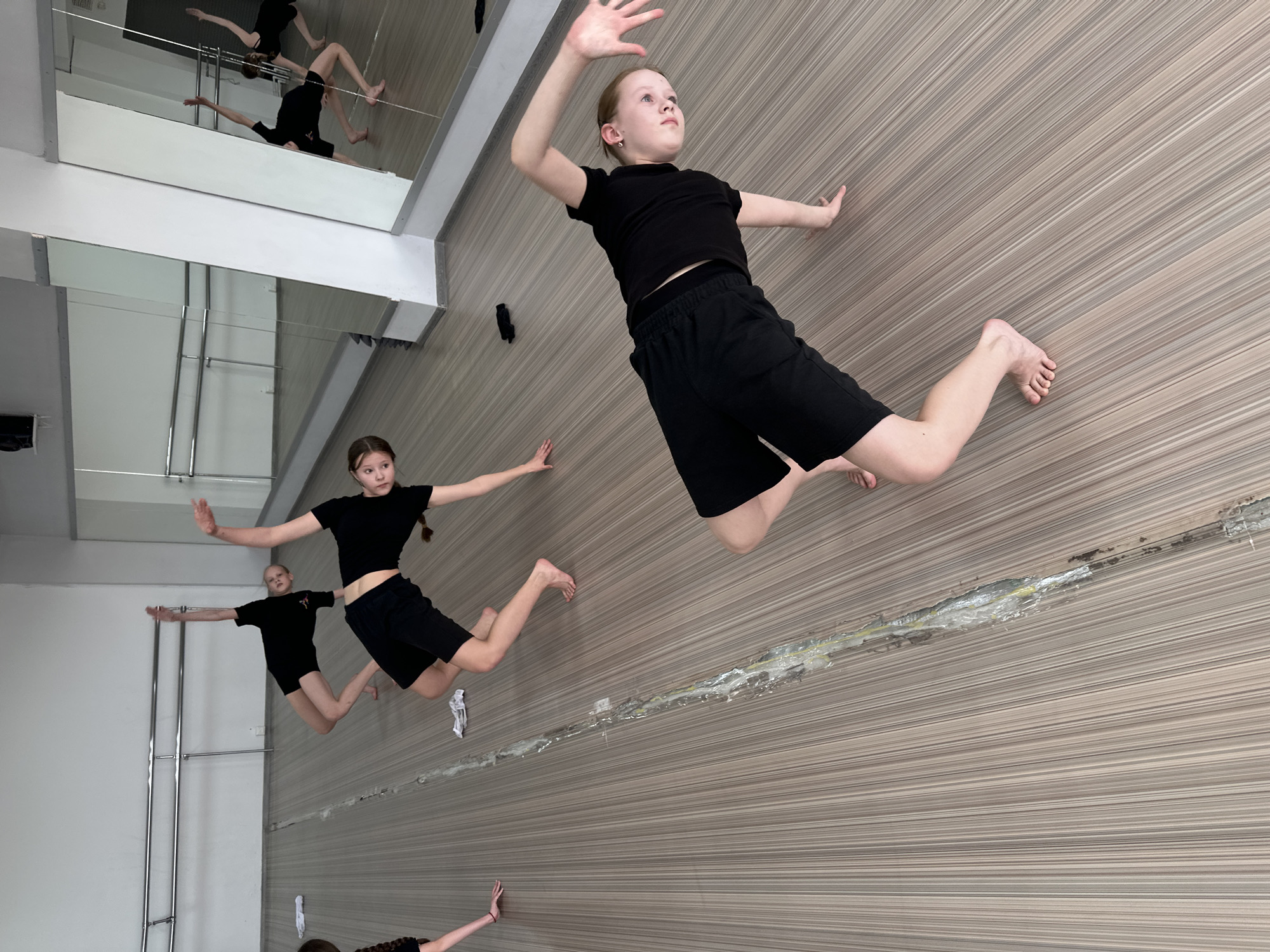 A photo taken indoors in what appears to be a dance studio in Votkinsk, Russia shows three young dancers in black shirts and shorts in mid-leap, mimicking a pose with arms extended and legs bent at the knees. The studio has a wall covered in horizontal striped paneling. There is a large mirror reflecting other dancers. There is white trim around the ceiling.