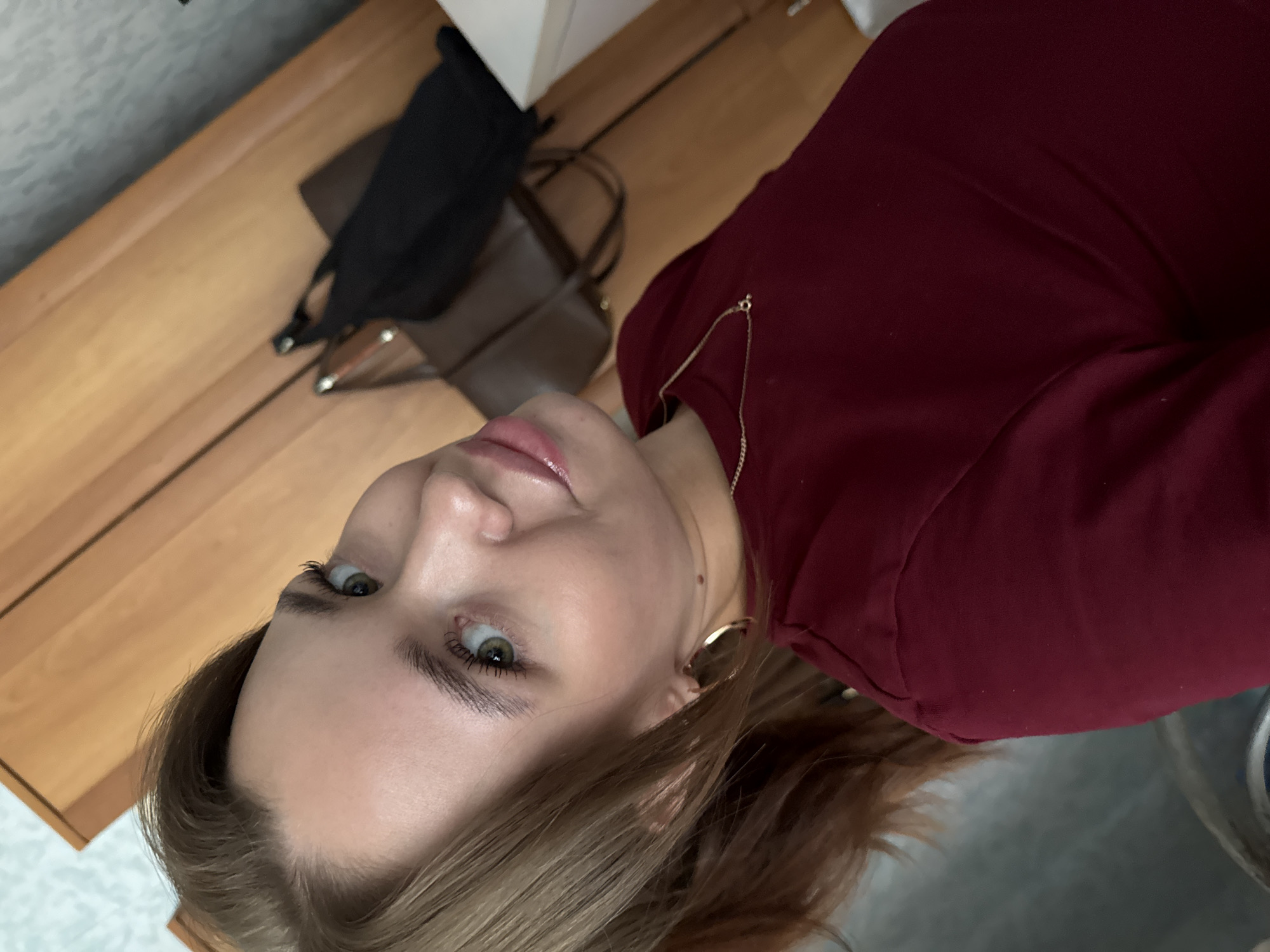 A young woman with light brown hair, green eyes, and light skin is taking a selfie. She is wearing a maroon long-sleeved shirt and a gold necklace. She has large hoop earrings. She is looking at the camera with a slight smile. In the background, there is a wooden wardrobe and a black bag. The setting appears to be a bedroom in an apartment in Chaikovsky, Russia.