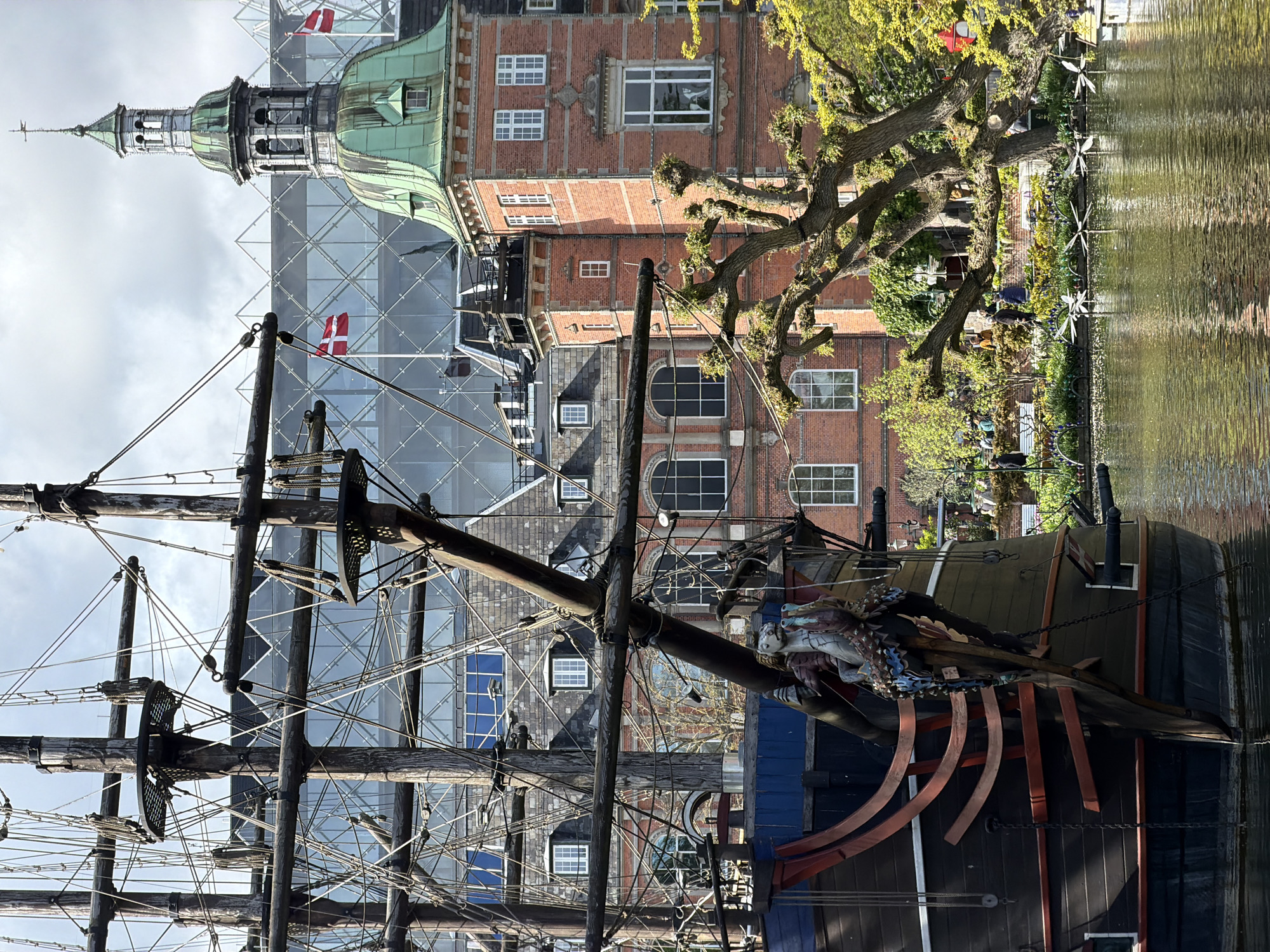 The image is taken at Nyhavn, Copenhagen, Denmark, and features a historical ship docked in the canal. The ship's mast and rigging fill the foreground, with a carved figurehead at the bow. Behind the ship are red brick buildings, some with scaffolding and Danish flags. On the right, trees line the canal. The sky is overcast.