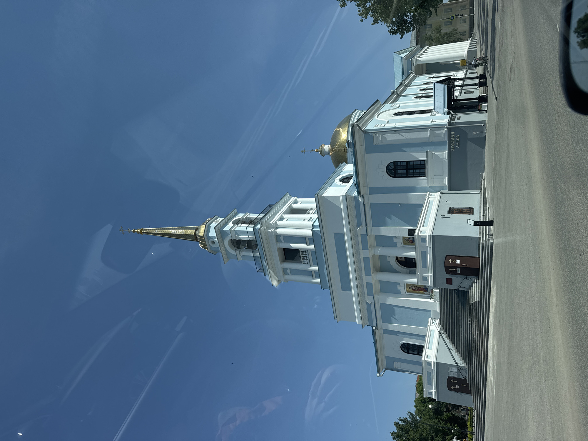 The image shows the Cathedral of the Annunciation in Votkinsk, Russia. The building is light blue and white with a gold dome and steeple. It is located on a street, and there are trees visible in the background. There are stairs leading up to the front entrance. There are two people on bicycles on the side of the church.