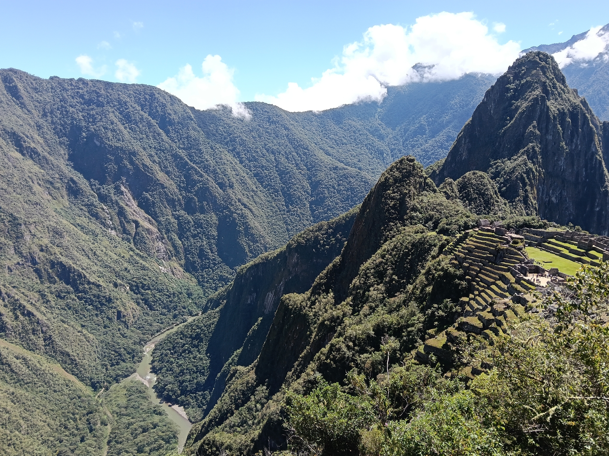 The image depicts Machu Picchu, Peru, a 15th-century Inca citadel. The site is located high in the Andes Mountains. The ruins consist of stone structures and terraces, partly overgrown with vegetation. A river flows through the valley below. Several mountains are visible, some covered with clouds. The sky is blue with patches of white clouds. Some people are walking in the area.