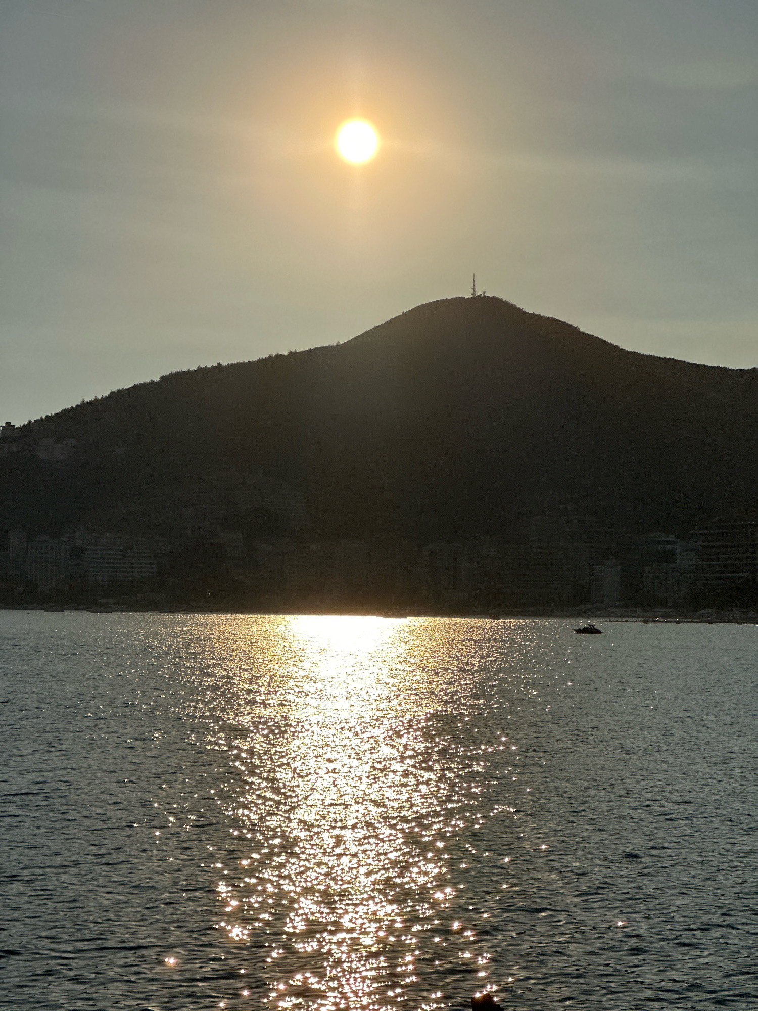 The image captures a scenic view of Budva, Montenegro, likely during sunset. The sun, a bright orb, is positioned high in the hazy sky, casting a golden glow across the water. A silhouetted mountain dominates the background, its slopes dotted with trees and buildings. The city's shoreline is visible at the base of the mountain, with numerous buildings forming a dense urban landscape. The sea in the foreground reflects the sunlight, creating a shimmering effect. A small boat is visible in the distance on the water. The overall atmosphere is serene and picturesque, showcasing the natural beauty and urban development of Budva.