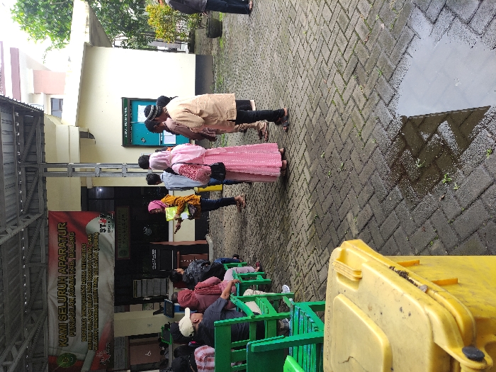 This media file captures an outdoor waiting area at the Religious Court of Malang Regency (Pengadilan Agama Kab. Malang) in Kepanjen, Indonesia. The ground is paved with wet brick tiles and features several puddles, indicating recent rain. The scene appears to be set during daytime under an overcast sky.

Numerous people are present, either standing or seated. A group of individuals, including men and women, some wearing headscarves and traditional attire, are standing in a line or queue near a dark green notice board on the building wall. Further into the foreground, other people are seated patiently on green plastic chairs arranged in rows, likely waiting for an administrative process or service. One person in the line is holding a distinctive pink item. In the lower right foreground, a yellow trash bin is partially visible.

A large banner hangs prominently on the left side of the frame, under a covered walkway. The text on this banner, in Indonesian, reads: "KAMI SELURUH APARATUR PENGADILAN AGAMA KAB. MALANG BERTEKAD MEWUJUDKAN ZONA INTEGRITAS MENUJU WILAYAH BEBAS KORUPSI (WBK) DAN WILAYAH BIROKRASI BERSIH DAN MELAYANI (WBM)". This translates to: "WE ALL APPARATUS OF THE RELIGIOUS COURT OF MALANG REGENCY ARE DETERMINED TO CREATE AN INTEGRITY ZONE TOWARDS A CORRUPTION-FREE REGION (WBK) AND A CLEAN AND SERVING BUREAUCRACY REGION (WBM)". This detail confirms the location and highlights the institution's stated commitment to good governance. The image portrays a typical scene at a public administrative office where citizens are engaged in various processes.