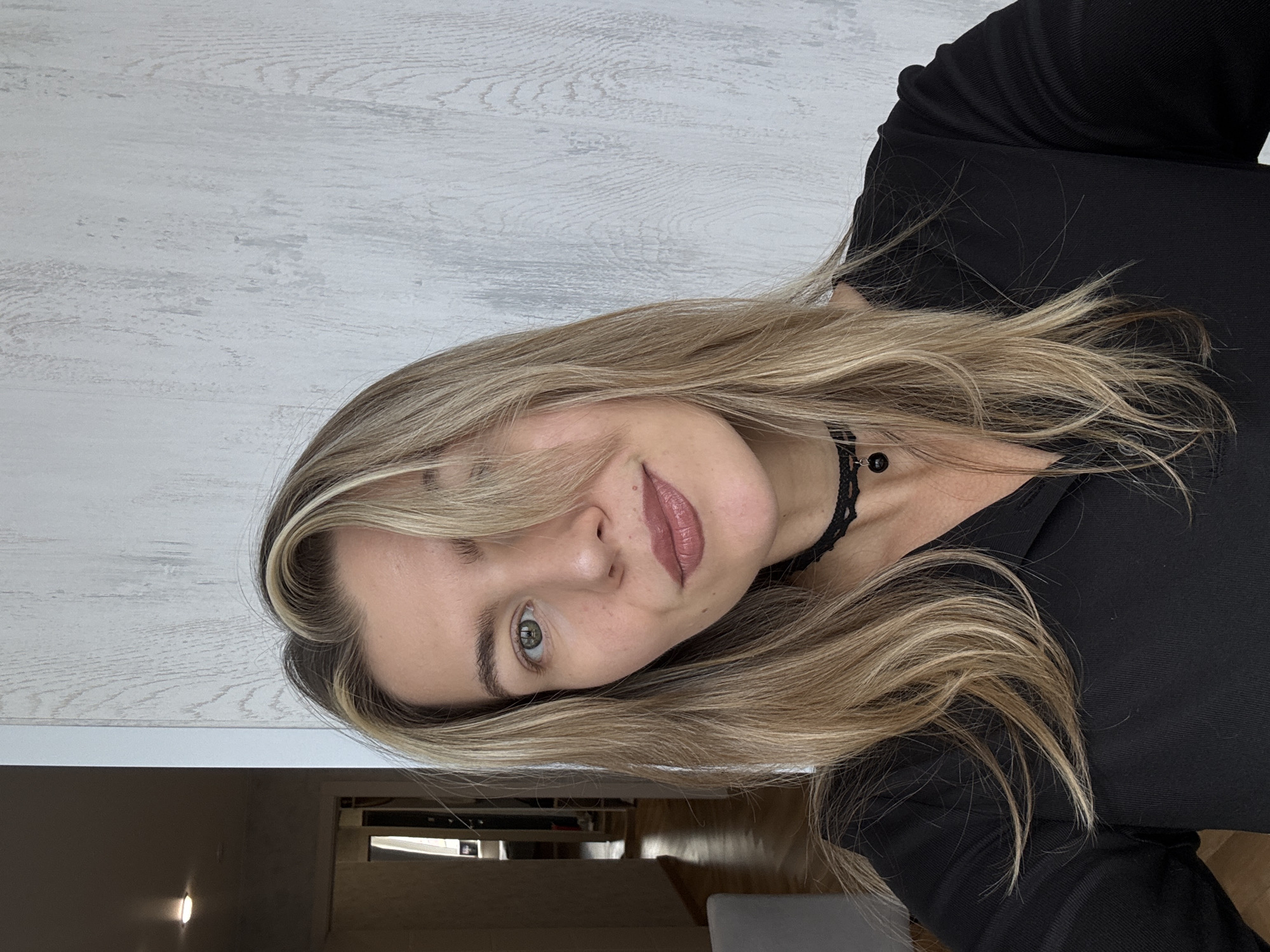 A woman with blonde hair and green eyes is posing for a portrait. She is wearing a black top and a black choker with a dark pendant. The background appears to be a wall with a wood-grain texture and a glimpse of a room with shelving and objects. The setting appears to be indoors. The location is Unknown, Unknown.