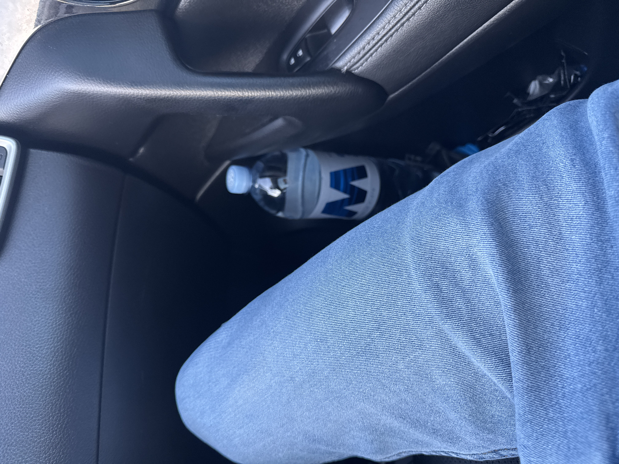 The image shows a close-up view of a person's legs wearing blue jeans, sitting in the passenger seat of a vehicle. A partially visible bottle of water with a blue "M" logo is placed in the door pocket. The interior of the car, including the dashboard and door panel, is visible. The scene is likely set in a residential area in Cojușna, Moldova, given the context of the photo.