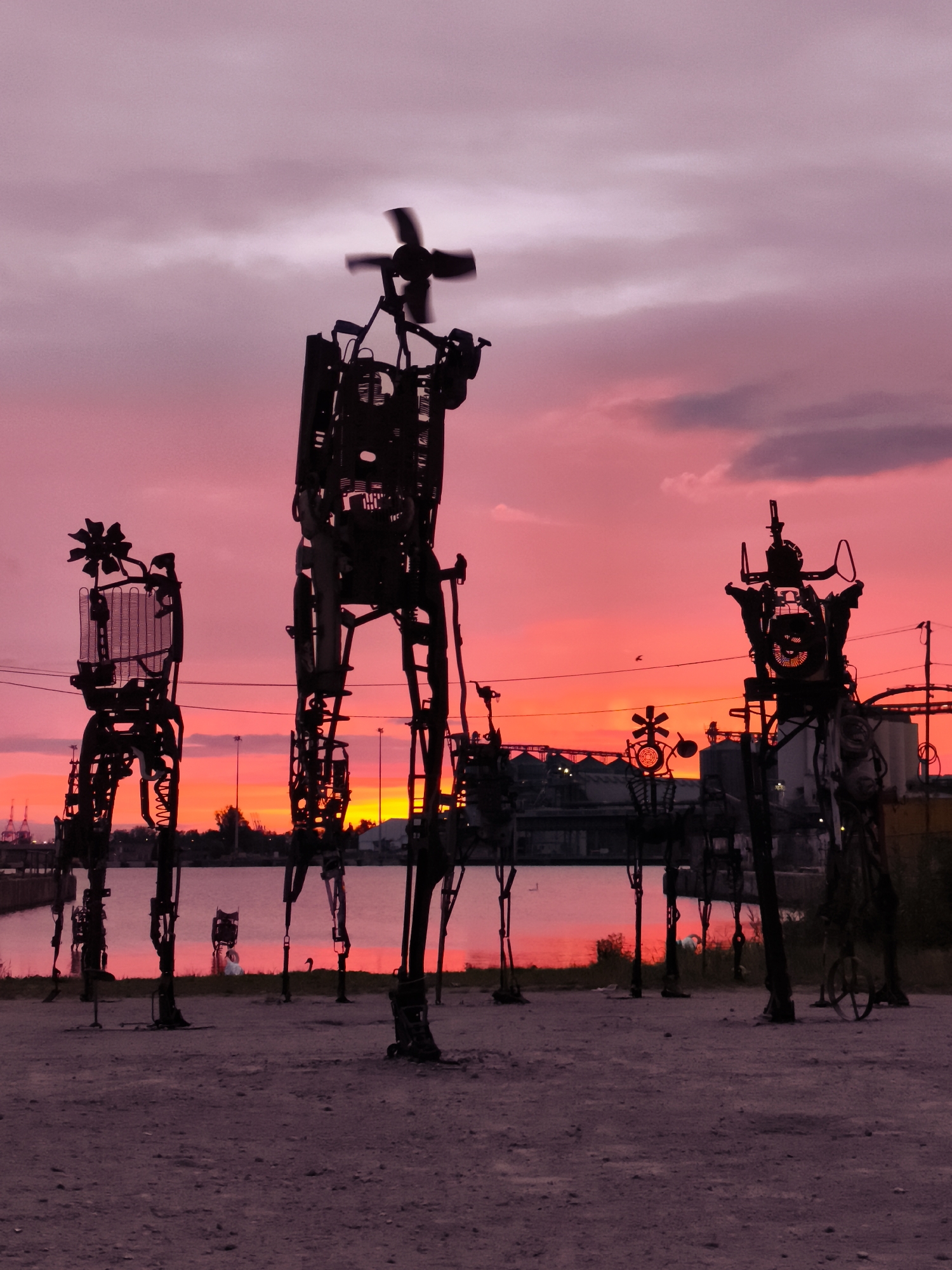 The image captures a striking sunset scene in the city of Gdańsk, Poland, specifically at the Monument to the Fallen Shipyard Workers. The sky is a vibrant gradient of pinks and oranges, with soft clouds adding texture. In the foreground, silhouetted against the colorful sky, are several large, abstract metal sculptures that resemble figures. These sculptures, made from scrap metal and industrial materials, are intricate and imposing. One of the most prominent figures in the center appears to have a propeller on its head. To the left, a smaller figure is topped with what looks like a flower or a decorative element. Further to the right, other metallic figures are visible, some with visible gears and mechanical components. The sculptures are positioned on a gravelly ground, with a body of water visible behind them, reflecting the sunset hues. In the background, along the water's edge, there are hints of industrial structures and lights, characteristic of a port city. The overall atmosphere is dramatic and artistic, highlighting the contrast between the industrial art and the natural beauty of the sunset.