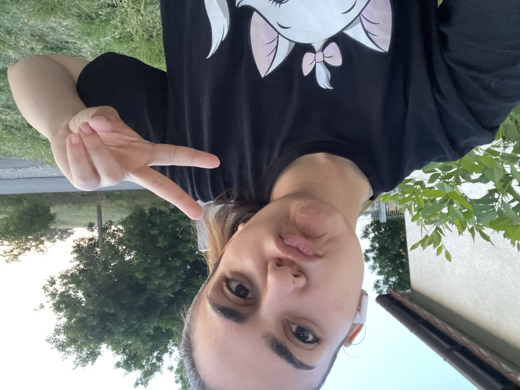 A young female, appearing to be in her early twenties, takes a selfie outdoors in Debelt, Bulgaria. She has dark hair pulled back, dark eyebrows, and dark eyes, looking directly at the camera with her lips pursed in a slight pout. A white wireless earbud is visible in her right ear. She wears a black short-sleeved t-shirt with a white cartoon cat, featuring a pink bow, printed on the chest. Her right hand (viewer's left) is raised, making a V-sign with her index and middle fingers.
The background on the left features a light-colored stucco or textured wall of a building with a dark brown gutter and roofline visible above. Green leafy foliage partially obscures the lower part of the building. To the right, dense green deciduous trees dominate the mid-ground, with a dark utility pole positioned vertically among them. An unpaved path or road is visible horizontally across the mid-ground. The far background reveals rolling grassy terrain with distant trees silhouetted against a pale, clear blue sky. The immediate foreground on the right consists of green grass and scattered weeds.