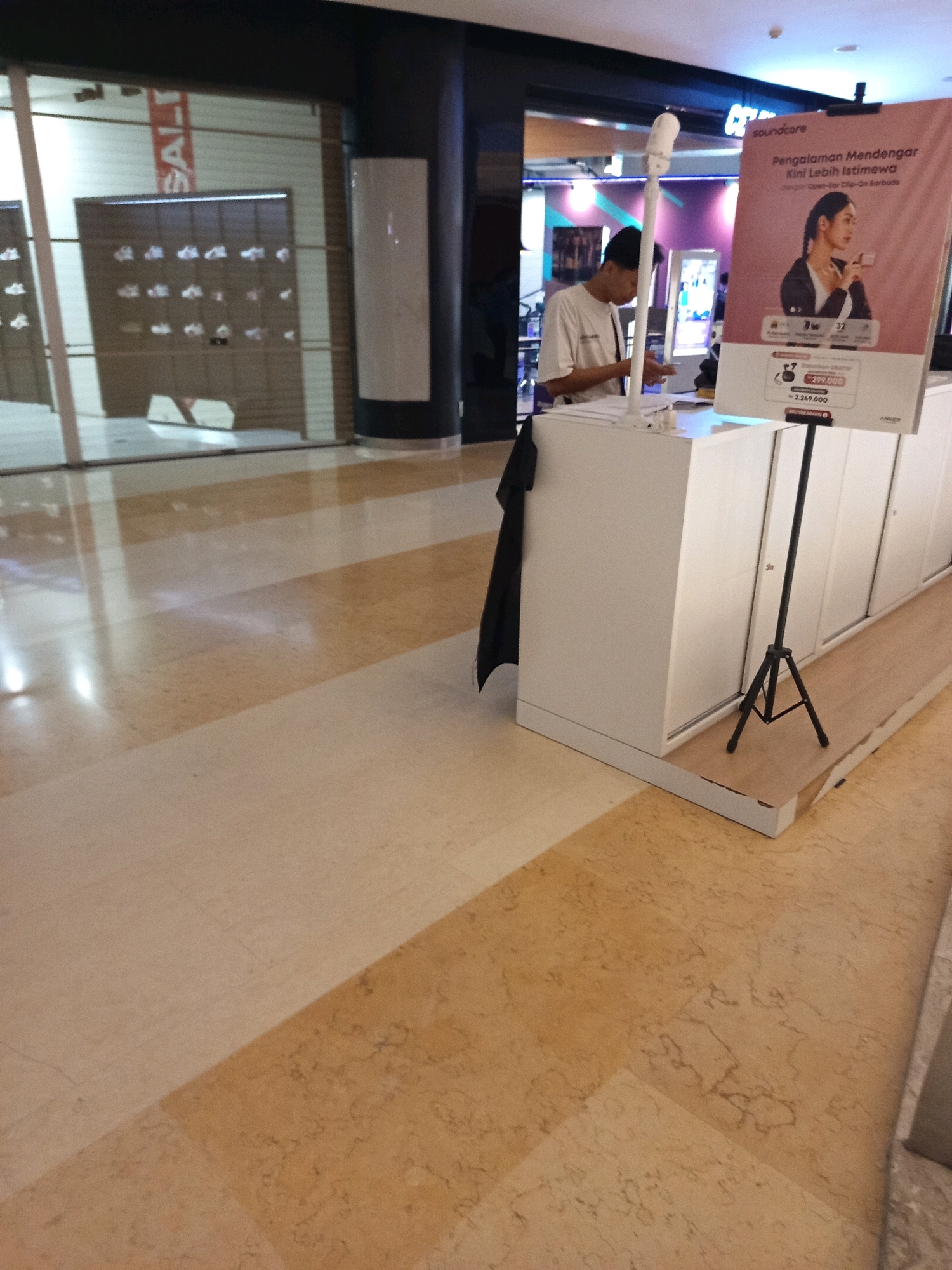 A man in a white t-shirt is standing behind a white counter in an indoor commercial space. He is looking down at something in his hands. To his right, there is a pink promotional banner for "soundcore" headphones. The banner features an image of a woman wearing headphones and text in Indonesian. To the right of the banner, there is a black tripod. The floor is tiled with a marble-like pattern of beige and brown. In the background, there is a storefront with shelves displaying items, and the word "SALT" is visible on a red banner. The overall environment appears to be a shopping mall or retail store. The location is identified as South Jakarta, Indonesia.
