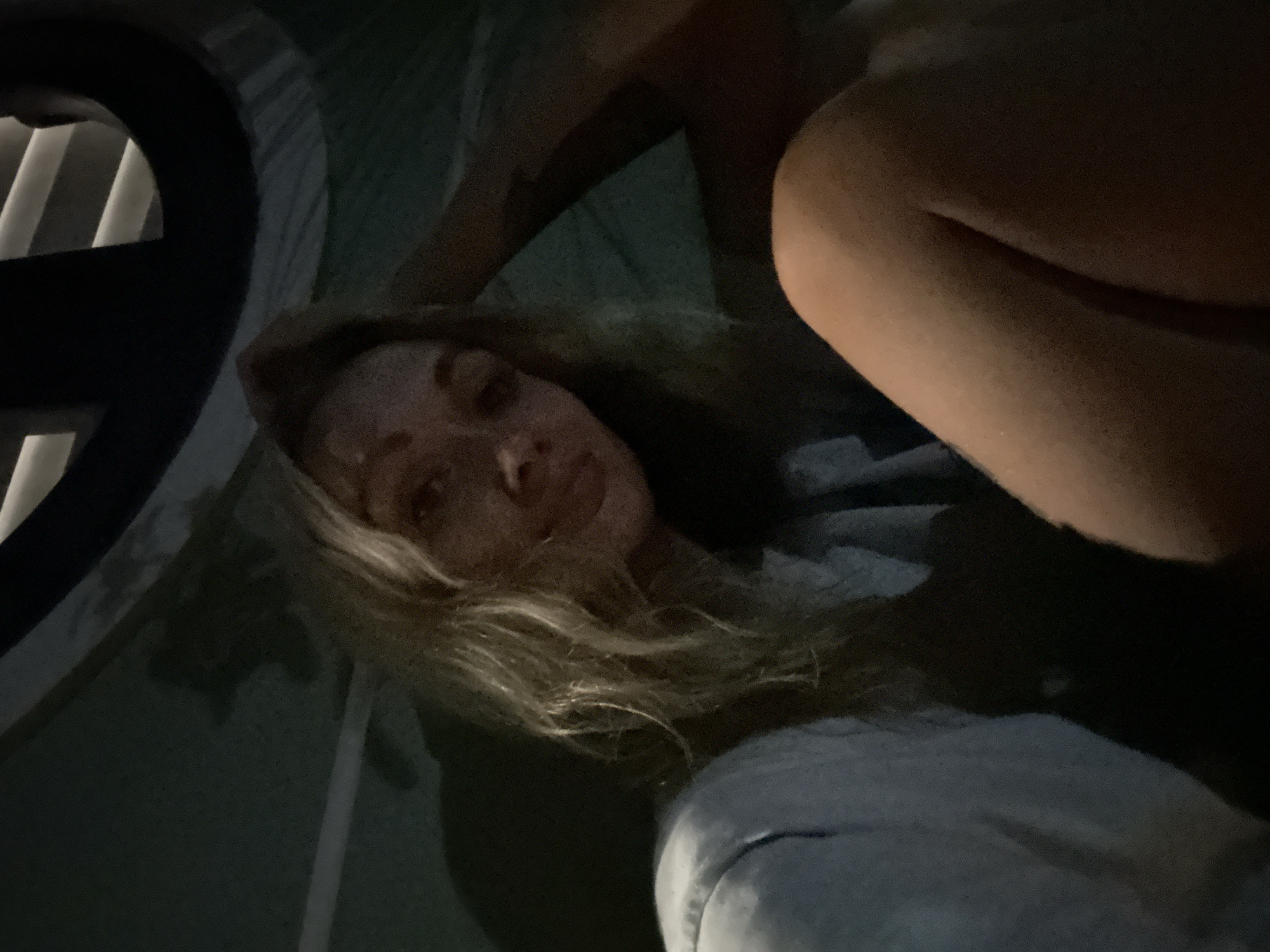A fair-skinned woman with light brown or blonde hair is lying inside a dark or dimly lit tent. The location is possibly Mũi Né, Vietnam, given the prompt's suggestion. The interior of the tent is green, and there is a black circular object with white horizontal lines visible on the left side of the frame. Her face is facing the camera, with a neutral expression. She is wearing a gray or light-colored shirt or sweater. Part of what appears to be a bare leg or arm is also in the frame in the upper right.