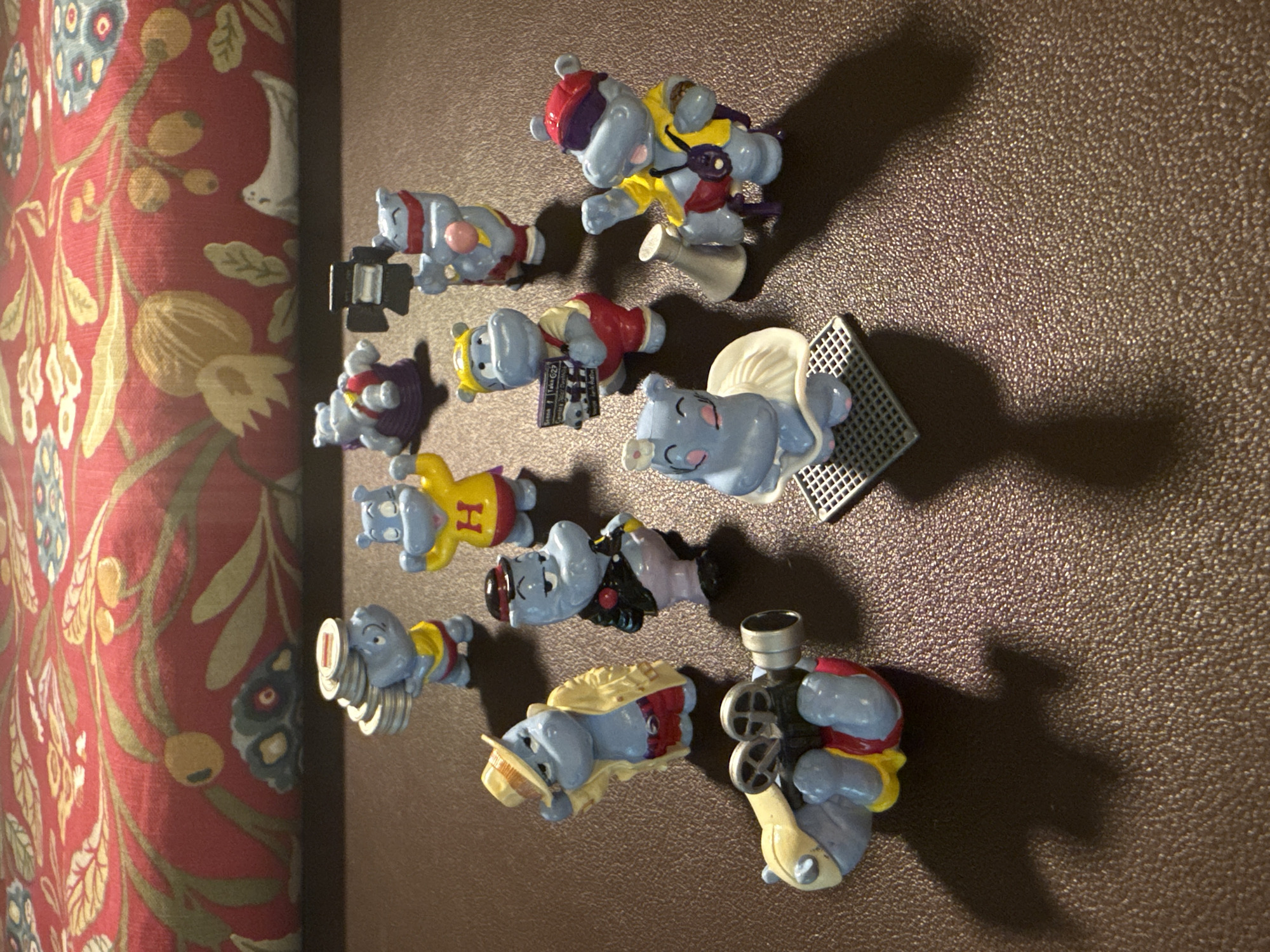 The image shows a collection of approximately twelve blue plastic hippopotamus figurines arranged on a brown textured surface. The figurines vary in pose and costume, suggesting they are part of a collectible set. Some are holding props like cameras or musical instruments. A patterned fabric, primarily red with floral motifs, is visible on the left side of the image. The setting does not appear to be Moscow, Russia, as there are no specific landmarks or elements suggesting that location.
