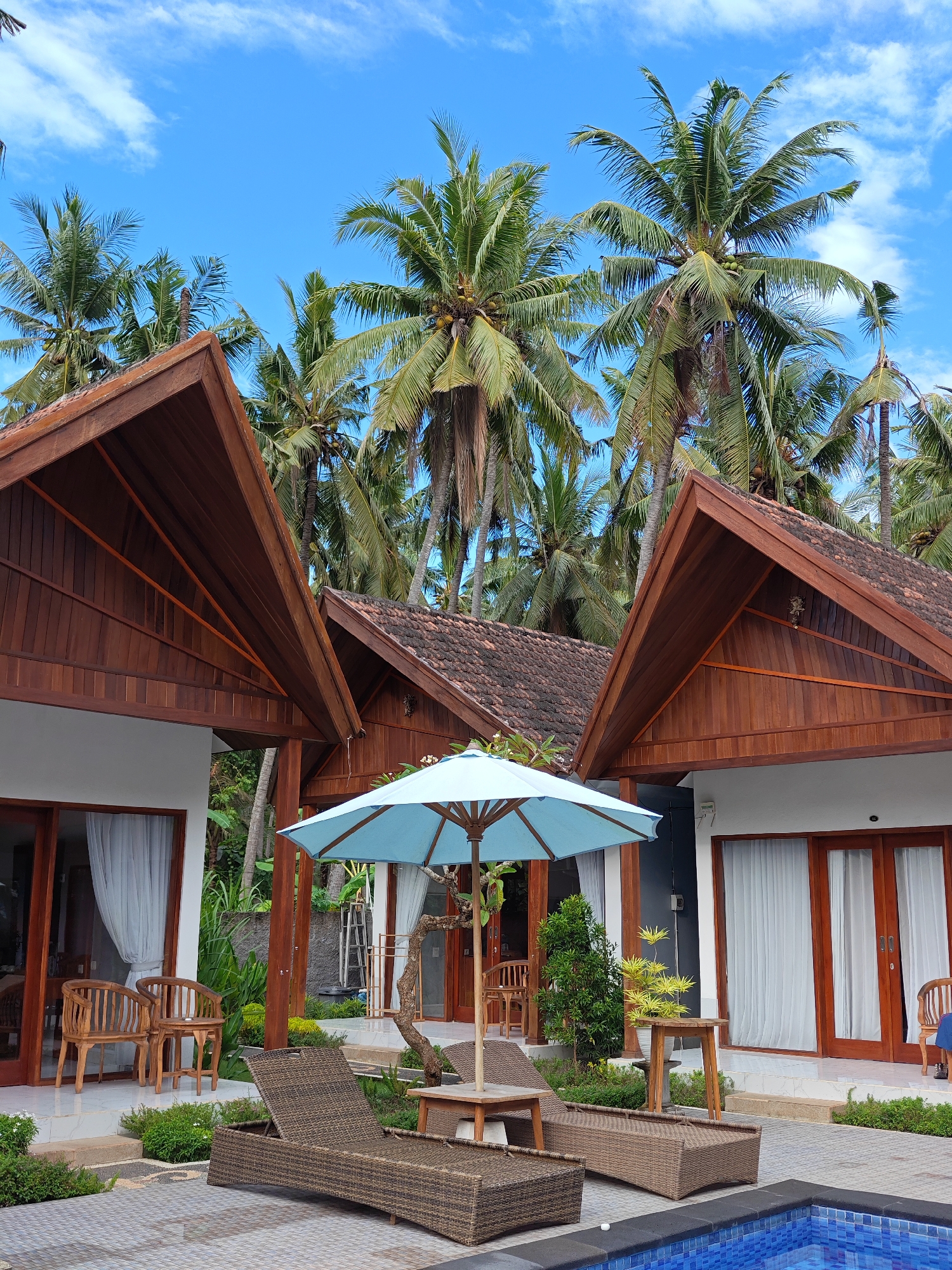 This is an outdoor shot of a resort area on Nusa Penida, Indonesia. Three villas are visible, featuring dark wooden gable roofs and white exterior walls. Large glass doors with light-colored curtains lead into each villa. Each villa has light-colored tiled patios.
In front of the villas, two wicker lounge chairs with dark brown cushions face a partially visible blue tiled pool. Between the lounge chairs and the middle villa, there is a light blue umbrella shading a small wooden table. The villas and lounge chairs are situated on a tiled patio area, with small plants bordering the edges. Lush palm trees fill the background, reaching up to a bright blue sky with scattered white clouds.
No people are directly visible.