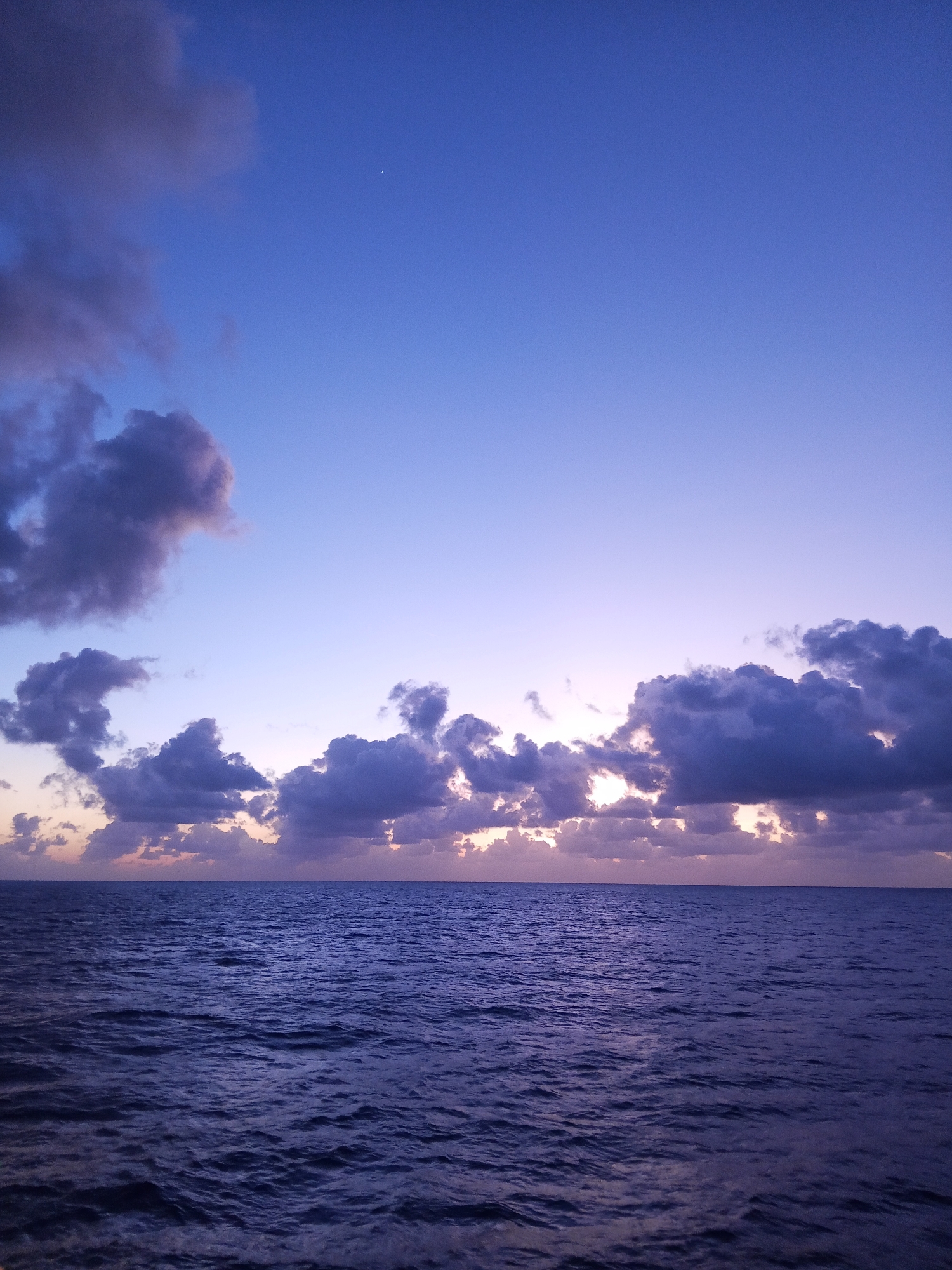 A vast expanse of dark blue ocean stretches to the horizon, its surface rippled by gentle waves. Above, the sky transitions from deep indigo at the top to a pale lavender near the horizon. Scattered across the sky are dramatic, dark purple clouds, some illuminated by unseen light sources that create glowing edges and internal highlights. A single, faint crescent moon is visible in the upper left quadrant of the sky. The scene depicts a tranquil, twilight setting over the open sea. The location is Unknown, Unknown.